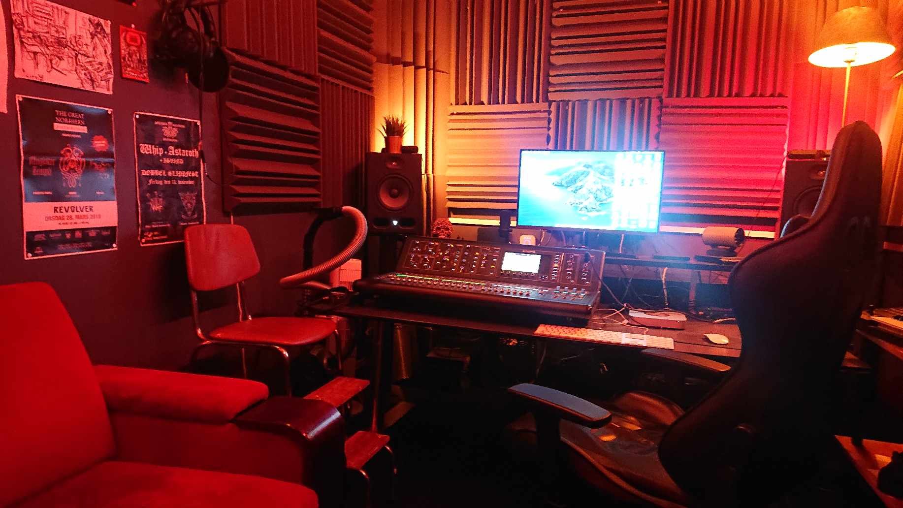 Recording studio with red lighting, featuring a mixing console on a desk, soundproofed walls, computer monitor, speakers, and chairs.