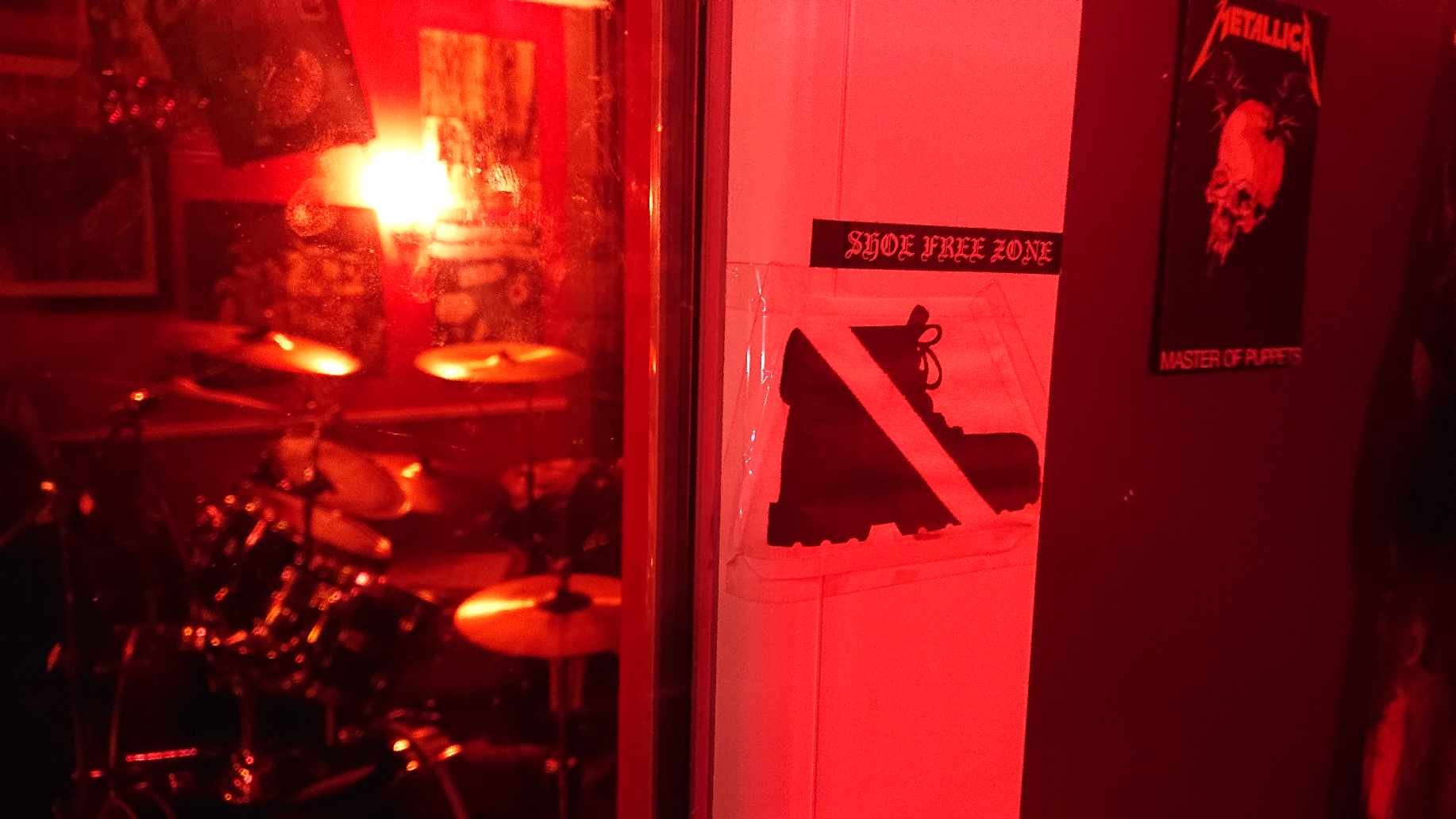 Room with red lighting, drum set, and signs indicating a shoe-free zone and a poster on the wall.