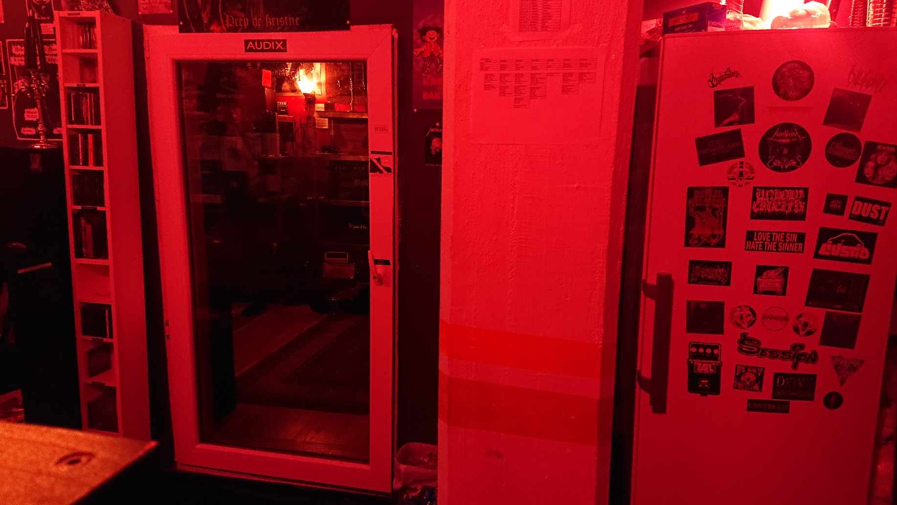 A room with red lighting featuring a door labeled 'AUDIX' and a refrigerator with multiple stickers. Shelves with various items are visible to the left.