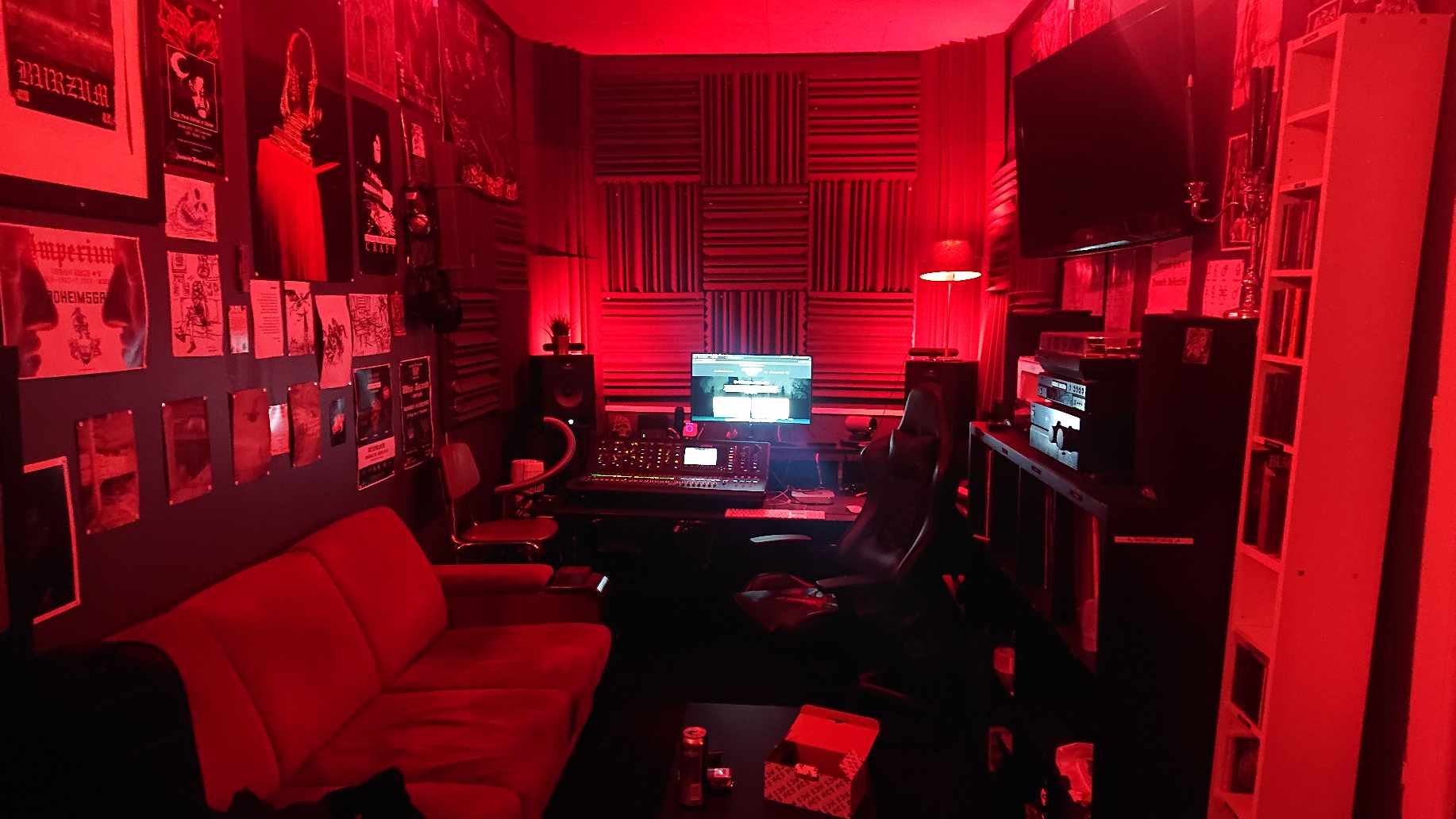 Recording studio with red lighting, soundproofing panels, mixing console, computer monitor, and wall posters.