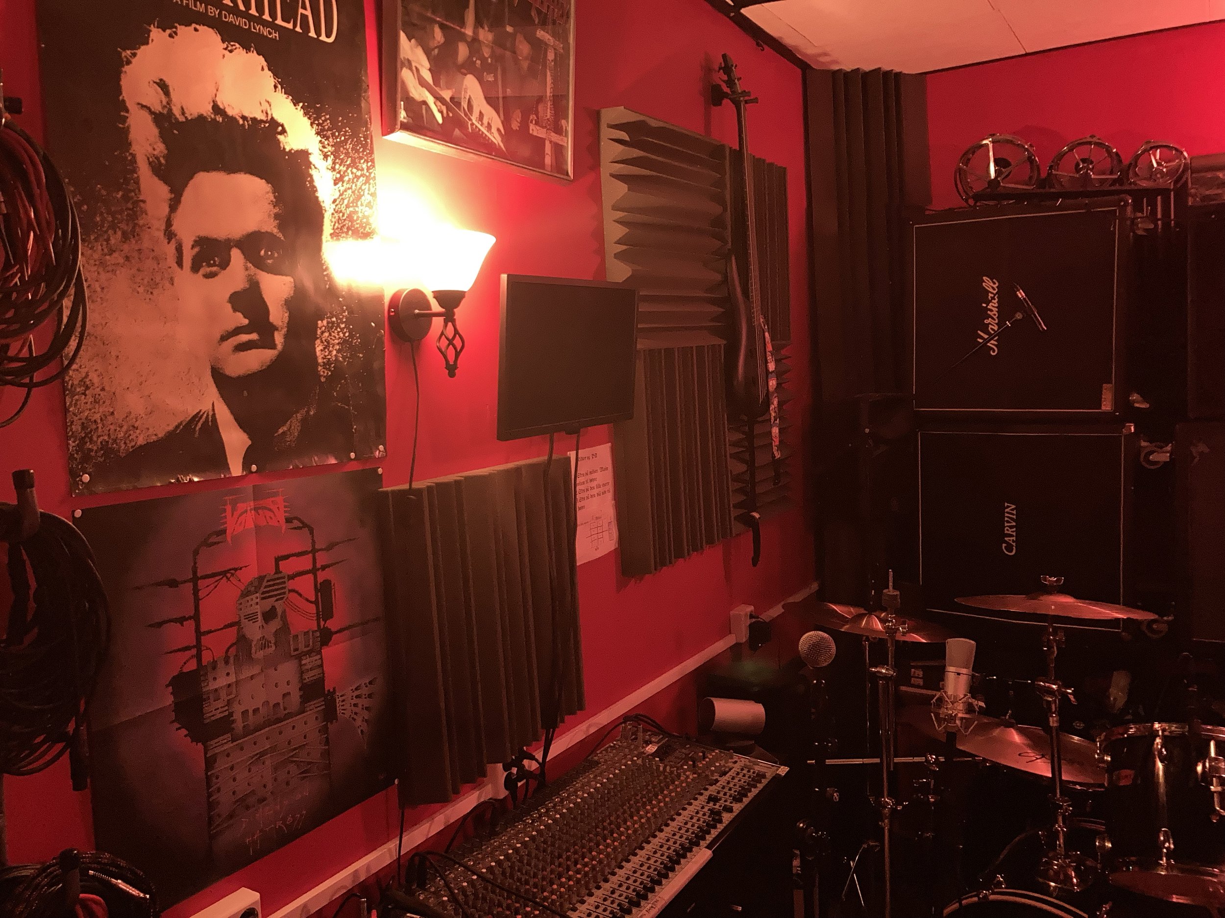 Music studio with red walls, featuring posters, a drum kit, mixing console, Marshall and Carvin amplifiers, and audio cables. Acoustic foam panels and microphones are also present.