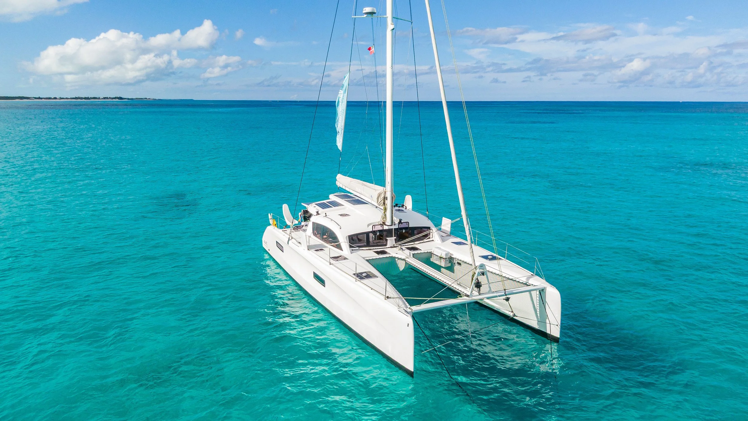 A white catamaran sailing boat with a tall mast floating on clear blue water under a partly cloudy sky, with land visible in the distance.