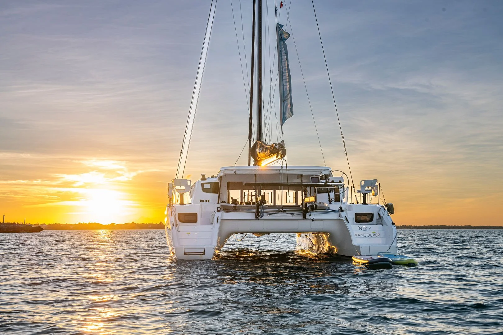 A white sailing catamaran with a tall mast floating on calm water during sunset, with a small inflatable dinghy attached at the stern and the sun setting behind the horizon.