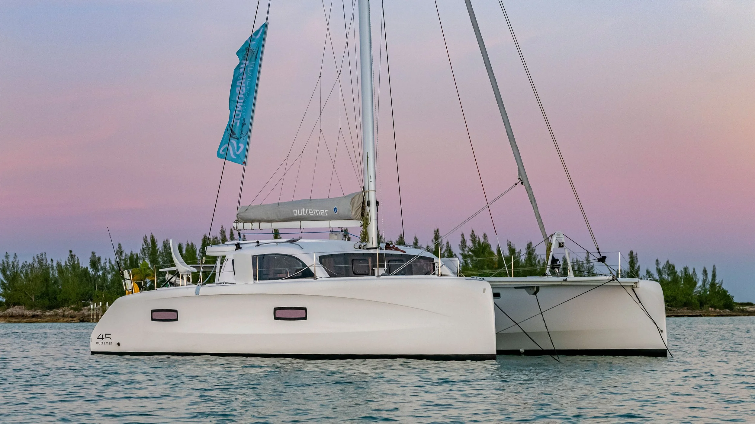 A white Outremer 45 catamaran sailing on calm water during sunset or sunrise with a pink sky and a treeline in the background.