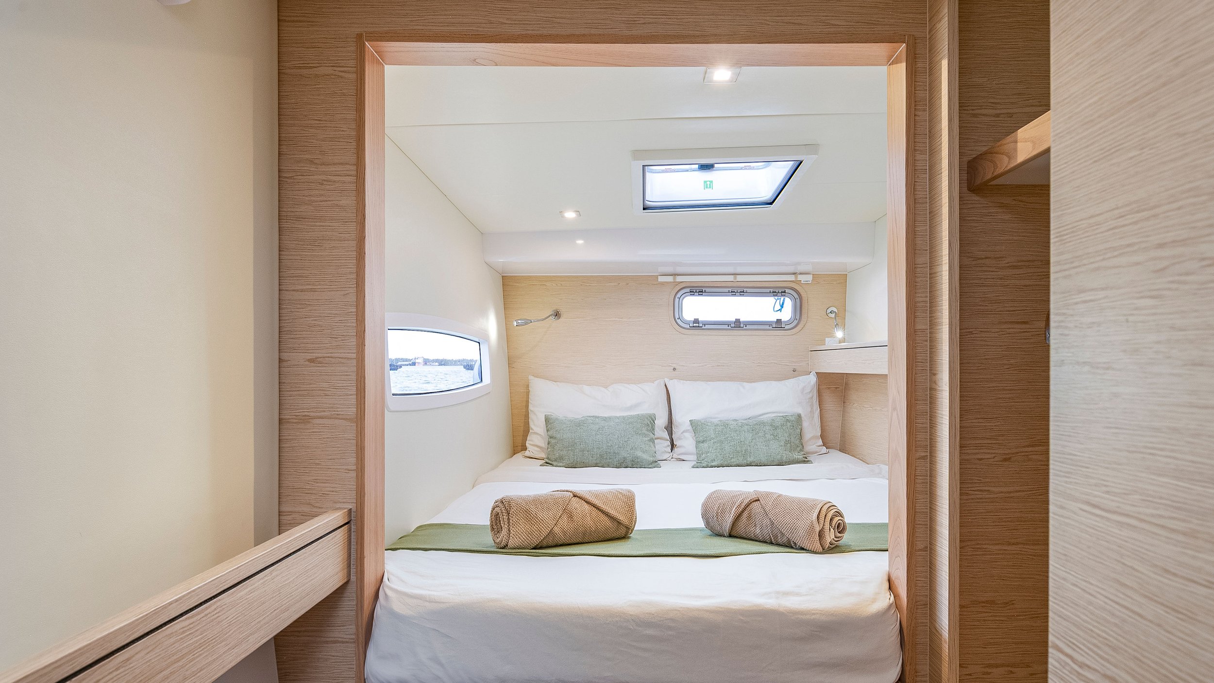 Interior view of a cozy bedroom with a bed covered in white linens, two rolled towels at the foot, and two green pillows. Small windows on either side of the bed reveal water outside, and a skylight is visible in the ceiling.