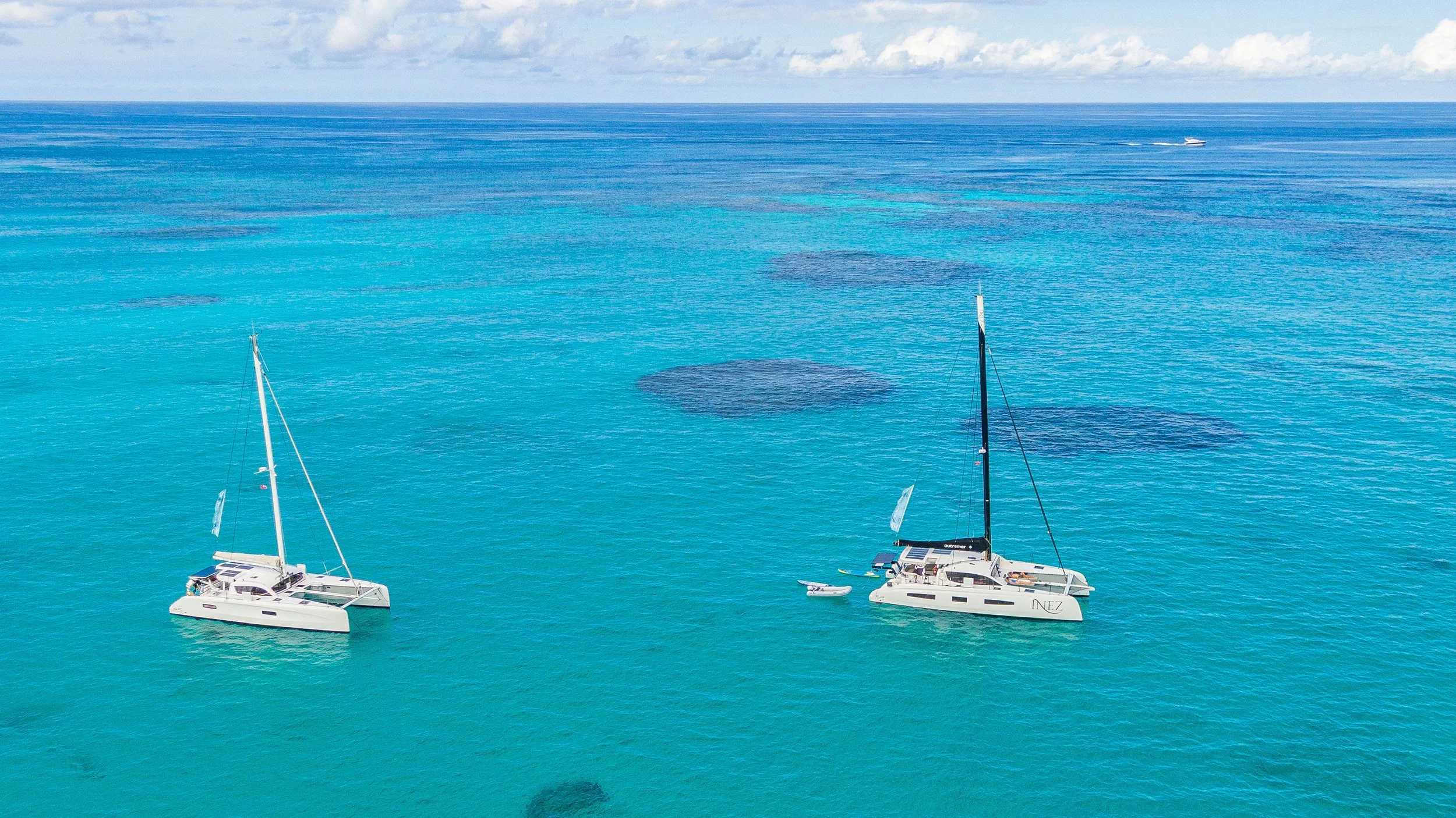 Two white sailboats anchored in clear blue ocean water near coral reefs, with a boat in the distance on the horizon.