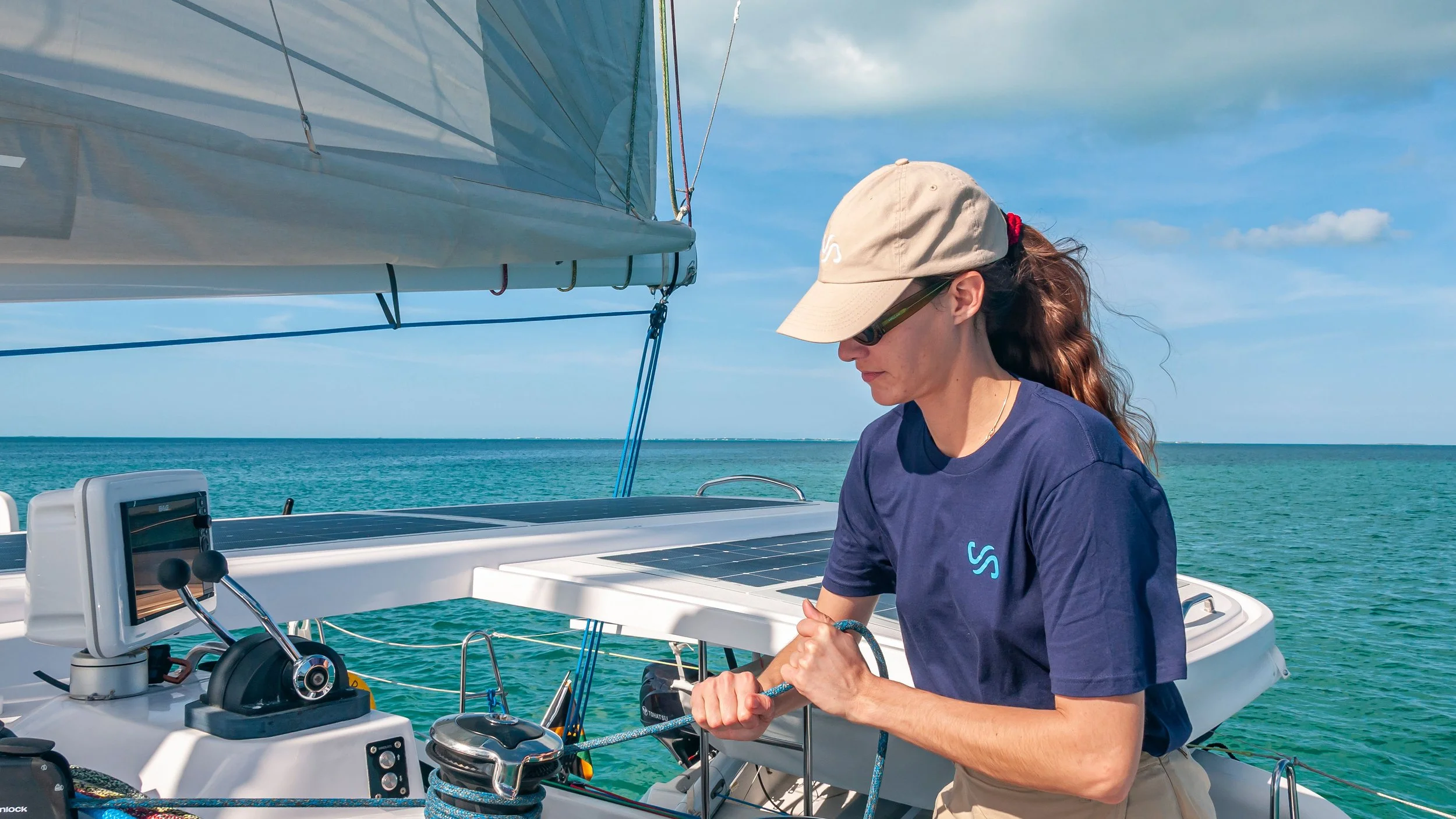 A woman on a sailboat adjusting the ropes, with the ocean and blue sky in the background.