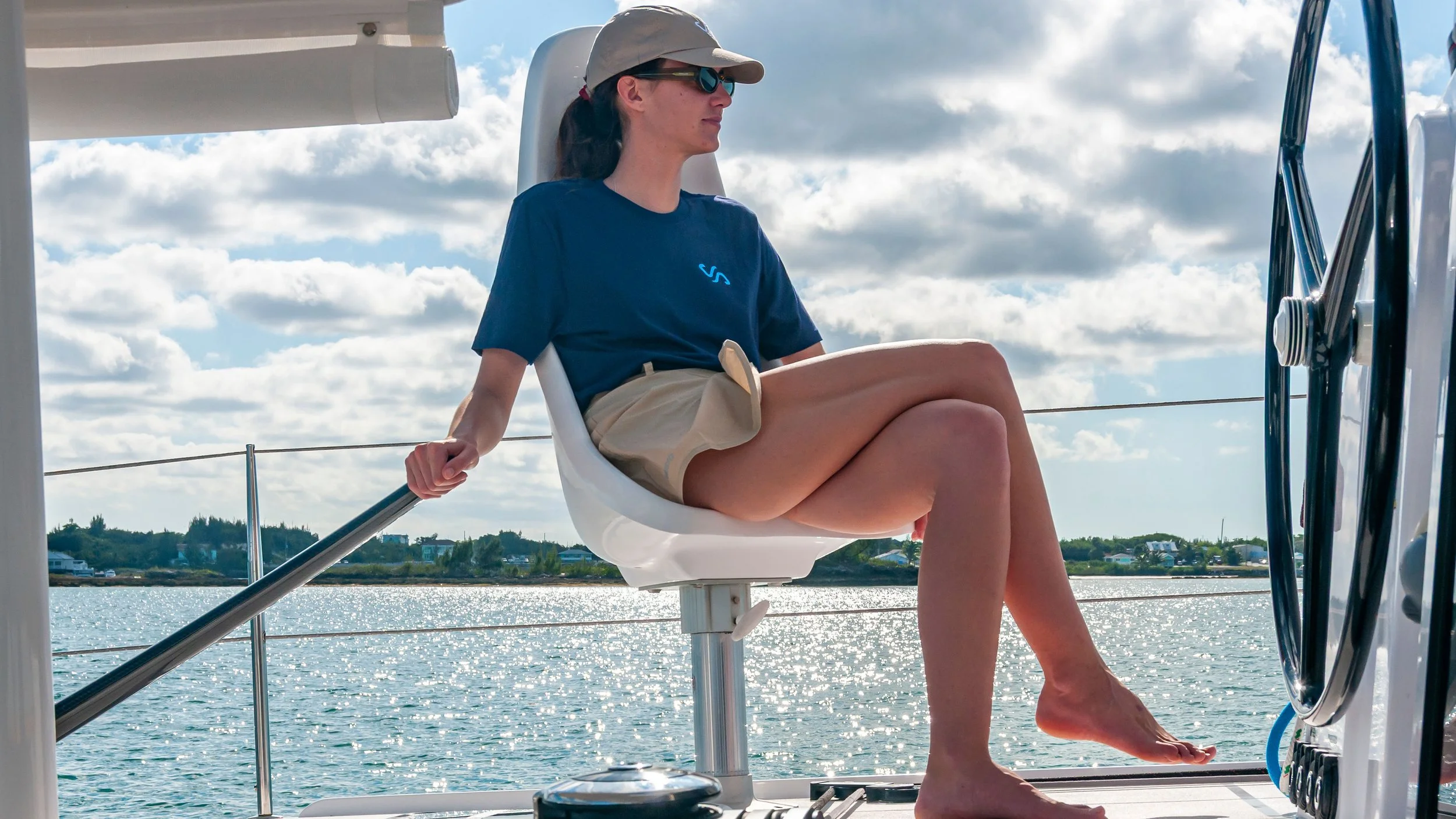 A woman sitting on a boat with water and a shoreline with buildings in the background, wearing sunglasses, a cap, a blue t-shirt, and beige shorts, holding onto a railing.