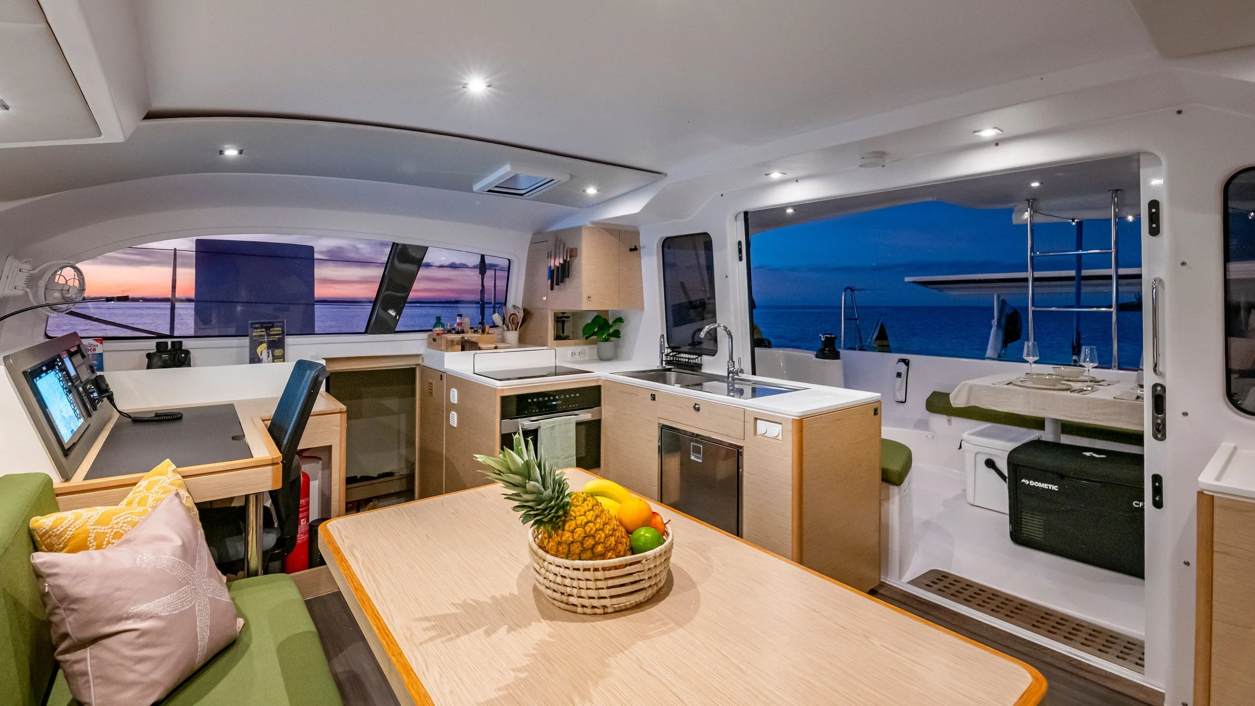 Interior of a modern yacht with a kitchen, dining area, and view of the ocean at sunset.
