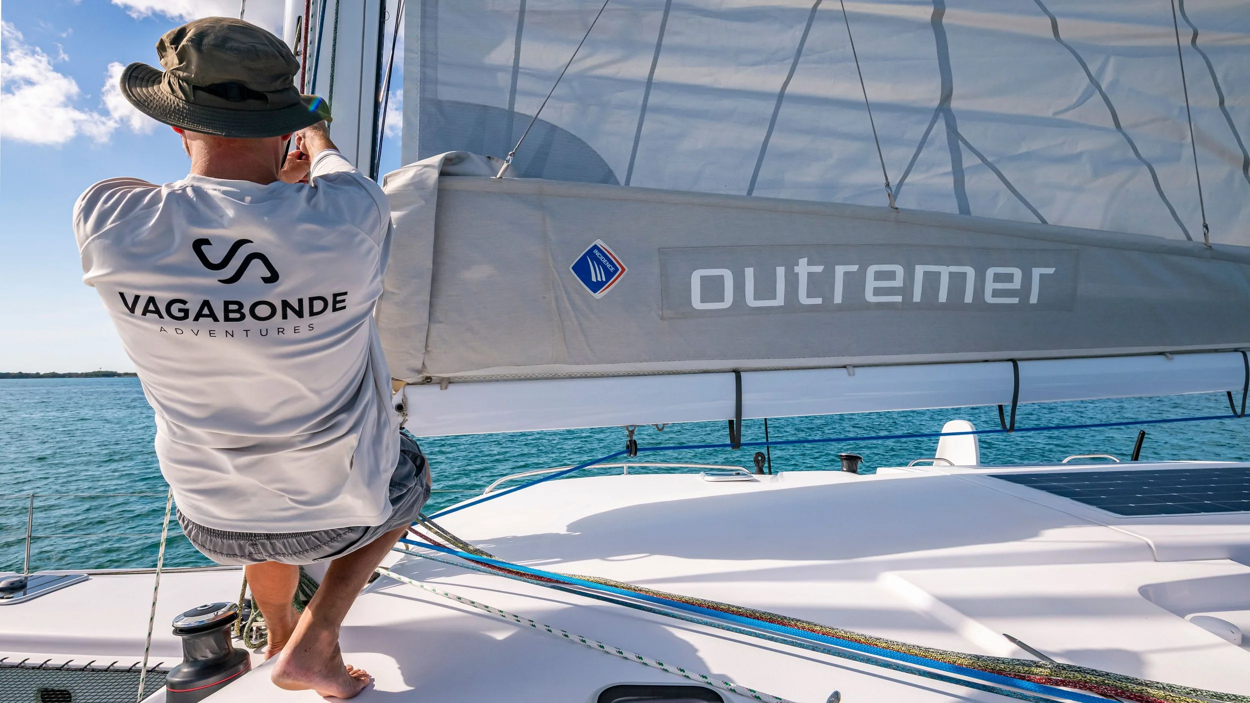 Person on a sailboat adjusting the sail, wearing a white shirt with 'VAGABONDE ADVENTURES' printed on the back, a hat, and shorts, with the ocean and a blue sky in the background.
