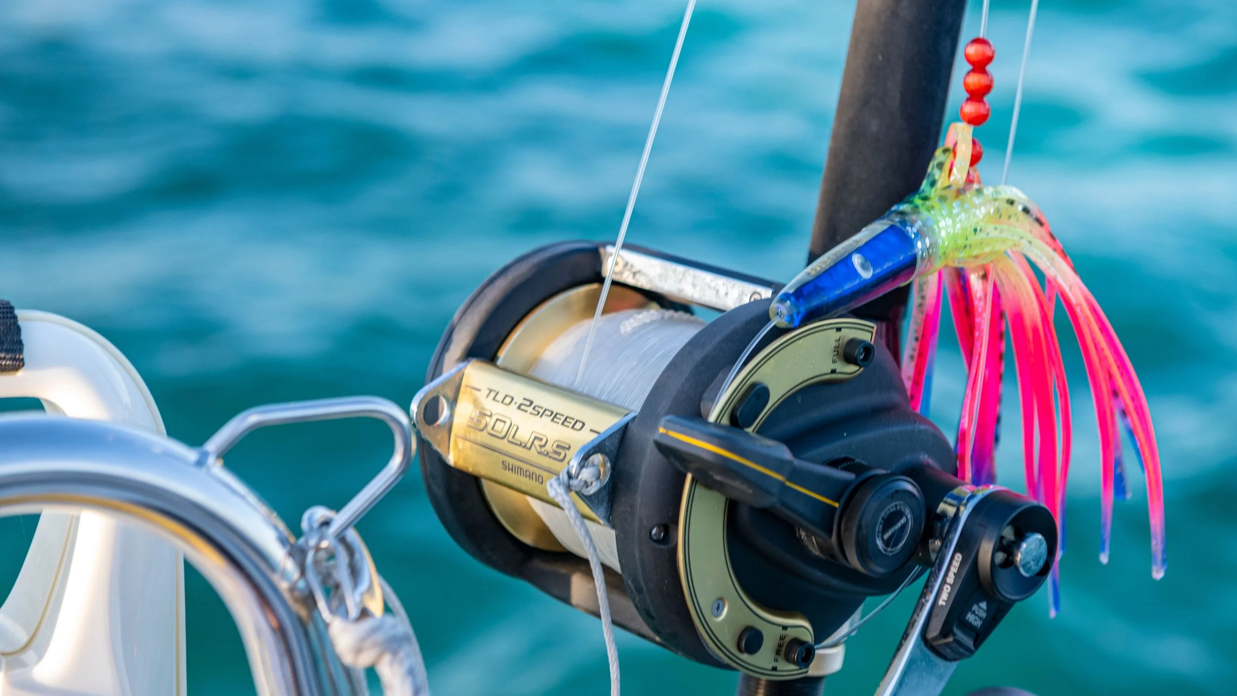 Close-up of a fishing reel mounted on a boat, with colorful artificial squid and pink and green fishing lures attached.
