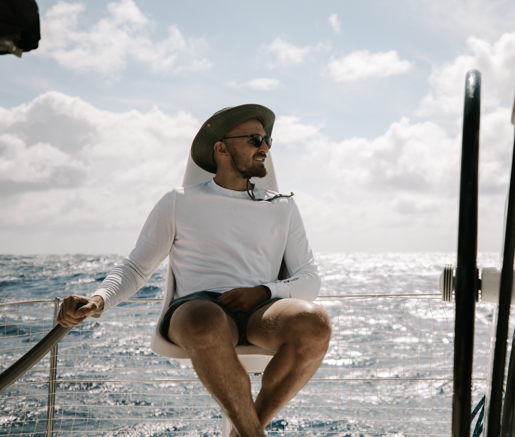 Man sitting on a boat with ocean in background, wearing a wide-brim hat, sunglasses, a white long-sleeve shirt, and shorts, enjoying sunny day on the water.