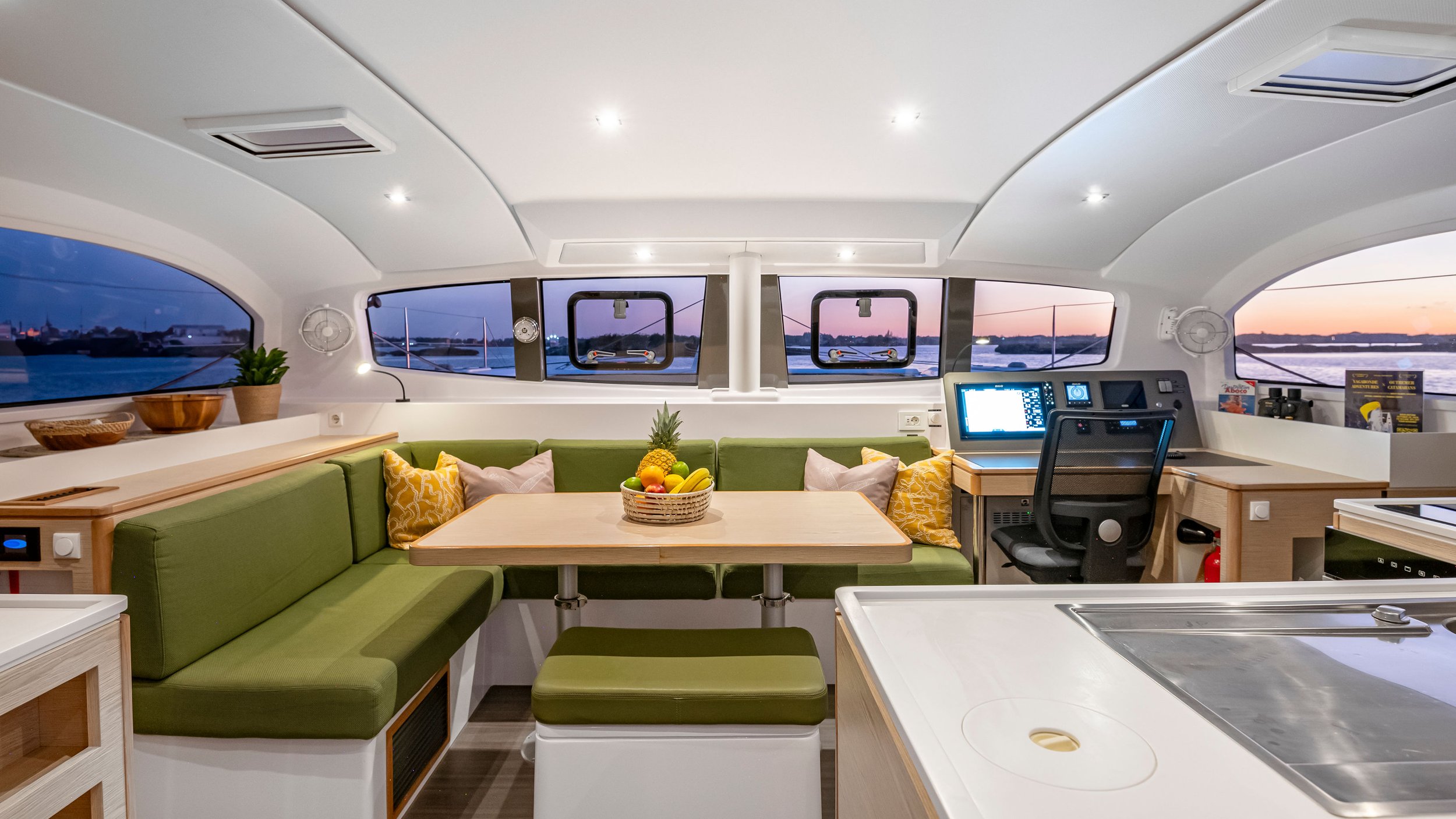 Interior of a boat with a dining area and navigation station, featuring a green sofa, a table with a fruit basket, and large windows showing a water view during sunset.