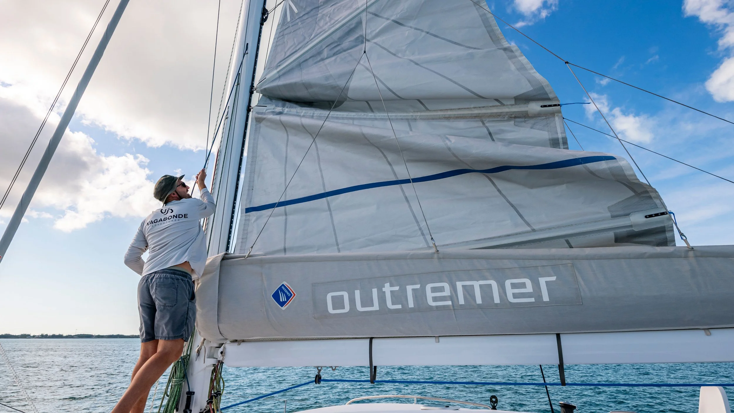 A man adjusting sails on a sailboat named Outremer on the water, with a blue sky and clouds in the background.