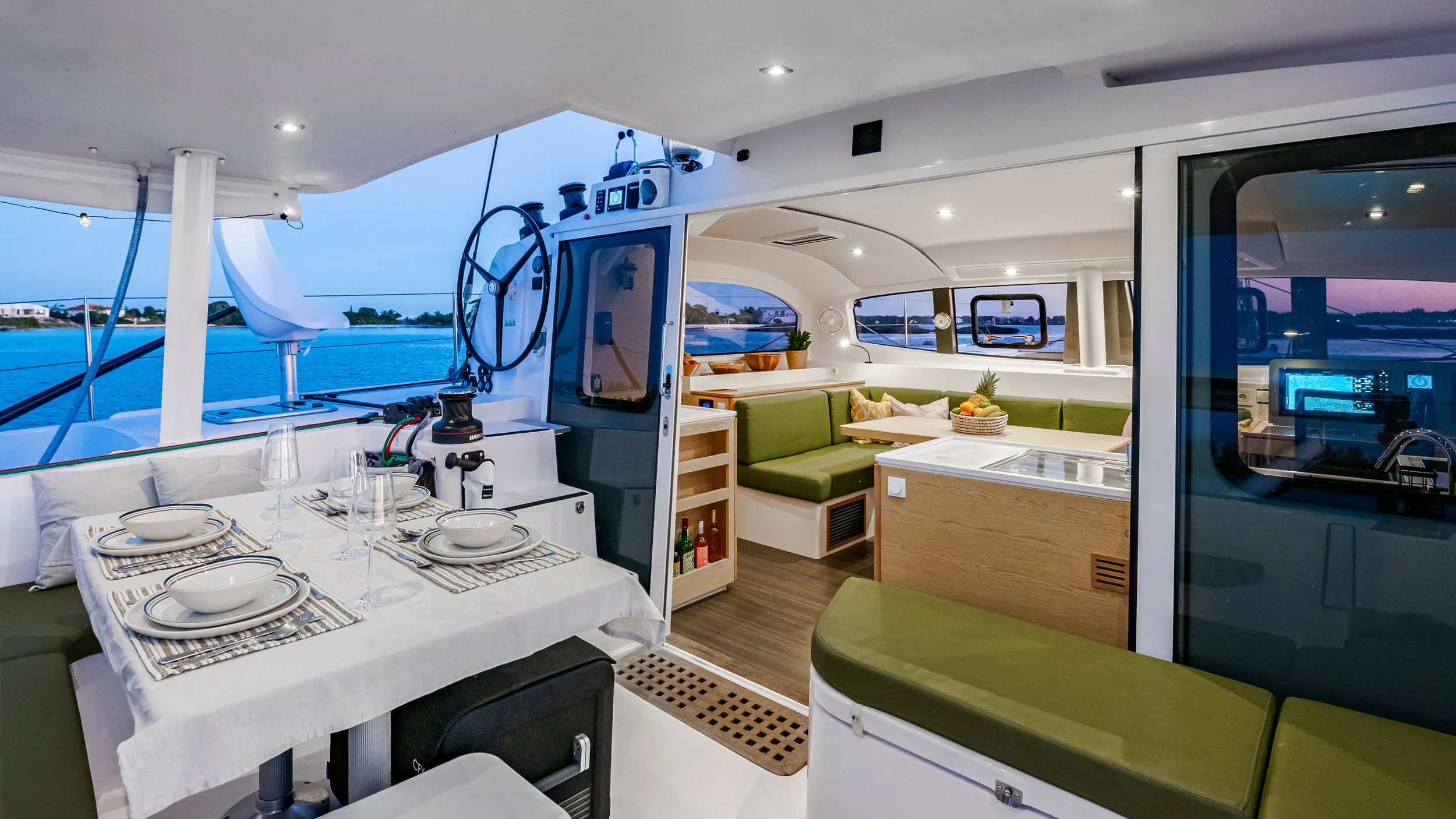 Interior of a yacht showing a dining table set with plates, glasses, and bowls, a compact kitchen area, and a lounge with green cushions and decorative pillows, with windows showing water and distant shoreline.