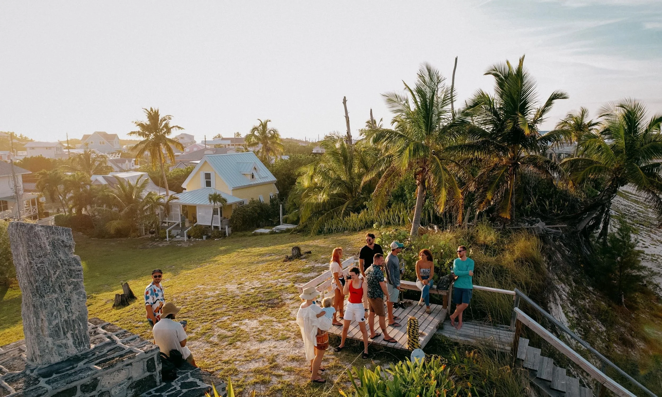 Group of people gathered on a wooden terrace overlooking a tropical landscape with palm trees and colorful houses during sunset.