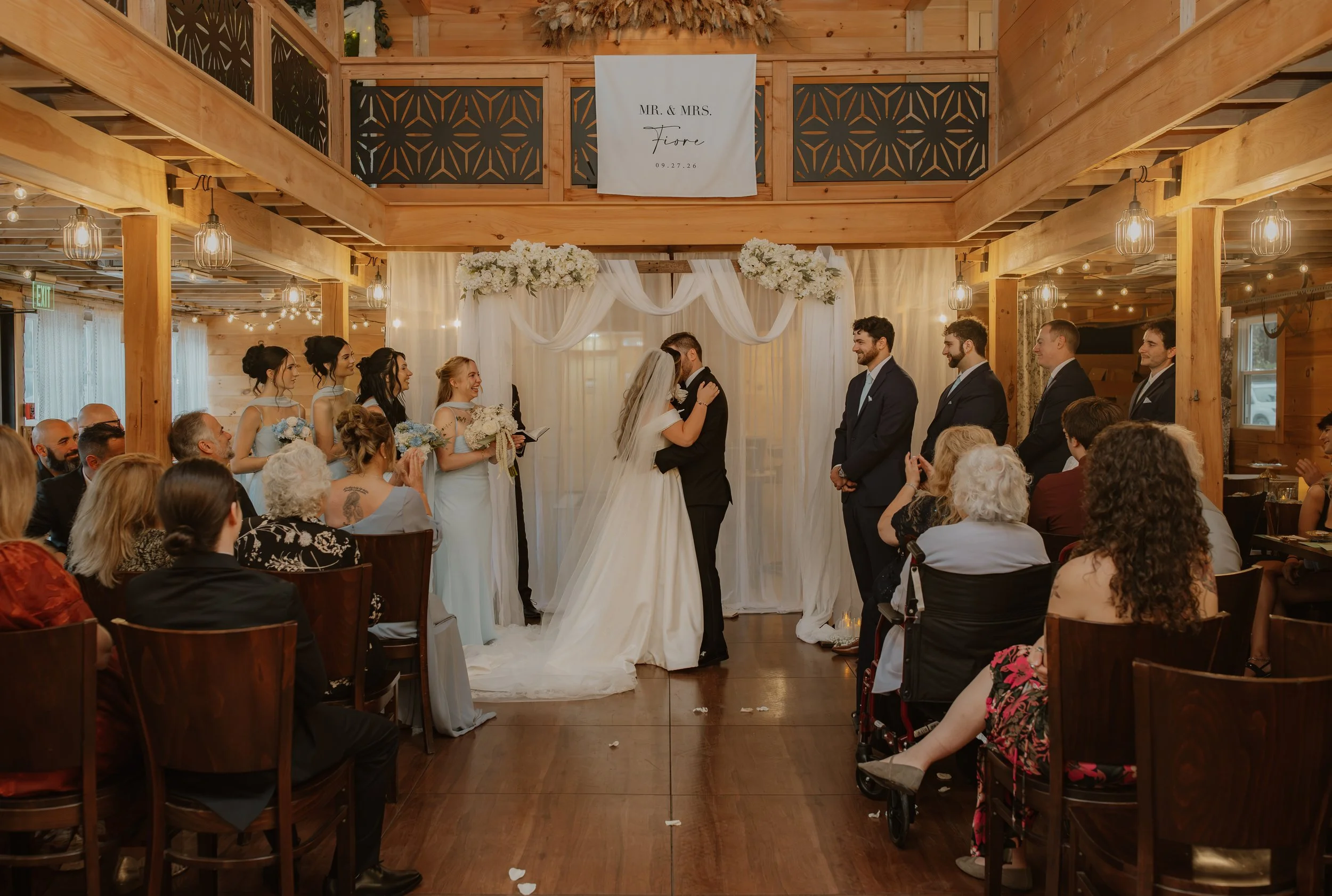 A wedding ceremony with a couple standing before an officiant inside a rustic wooden venue. The bride is in a white lace wedding dress with a veil, and the groom is in a light-colored suit. The officiant is reading from a black tablet. The ceremony altar is decorated with dried pampas grass and neutral-toned drapery, with warm ambient lighting.