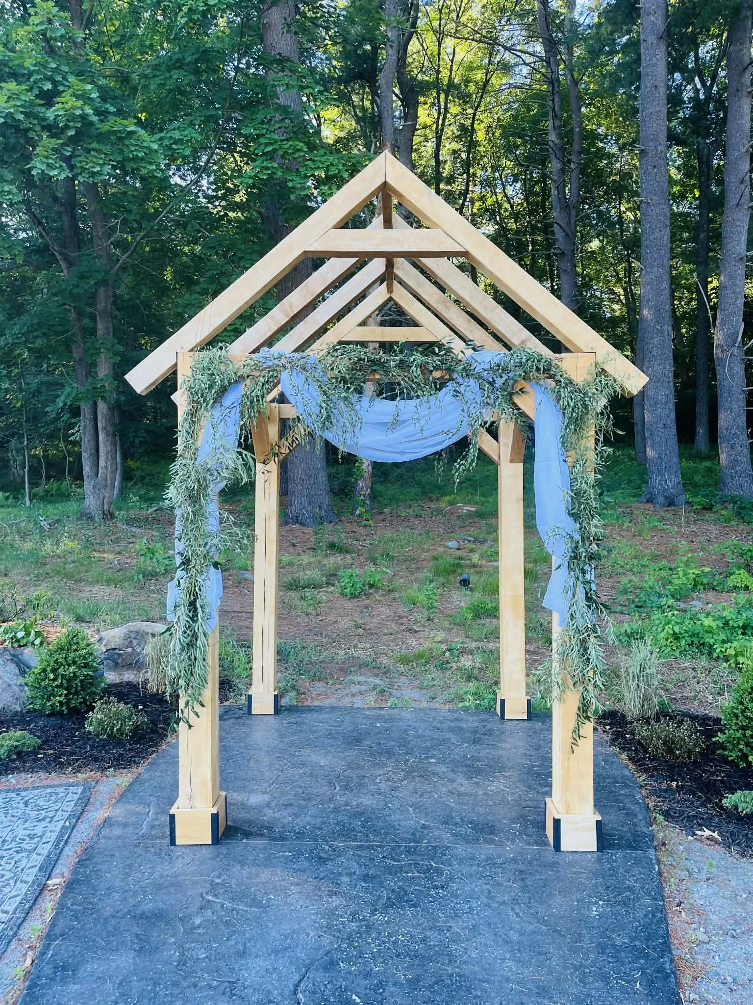 Outdoor wedding ceremony with guests seated on benches, a bride and groom standing in front of an officiant, surrounded by tall trees, clear sky, and bright sunlight.