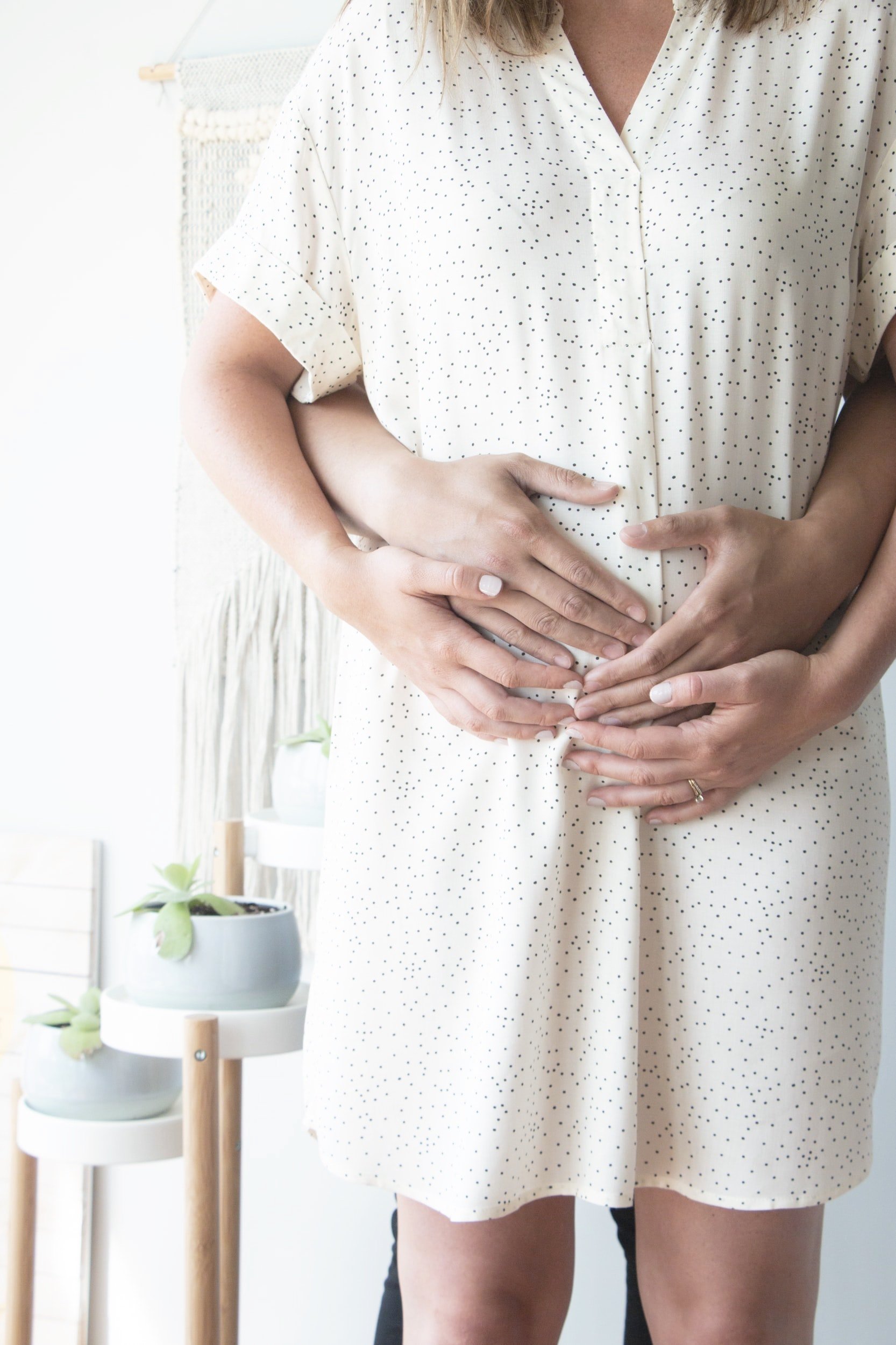 A pregnant woman wearing a white dress with black polka dots, holding her belly with both hands, standing indoors near a small white table with potted plants.
