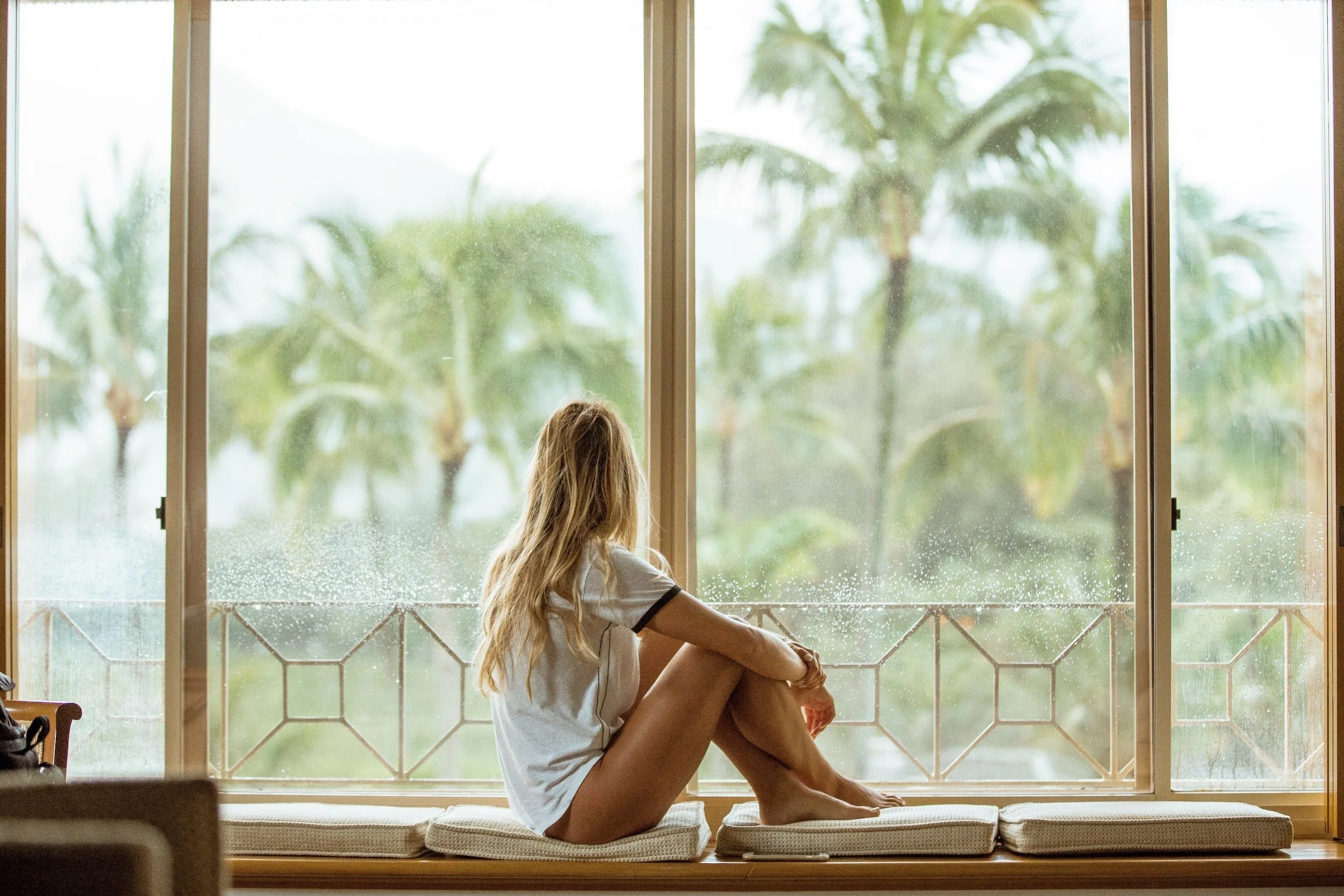 A young woman with long blonde hair sitting on a window seat with pajama shorts and a white T-shirt, looking out at a rain-soaked window and lush green palm trees outside.