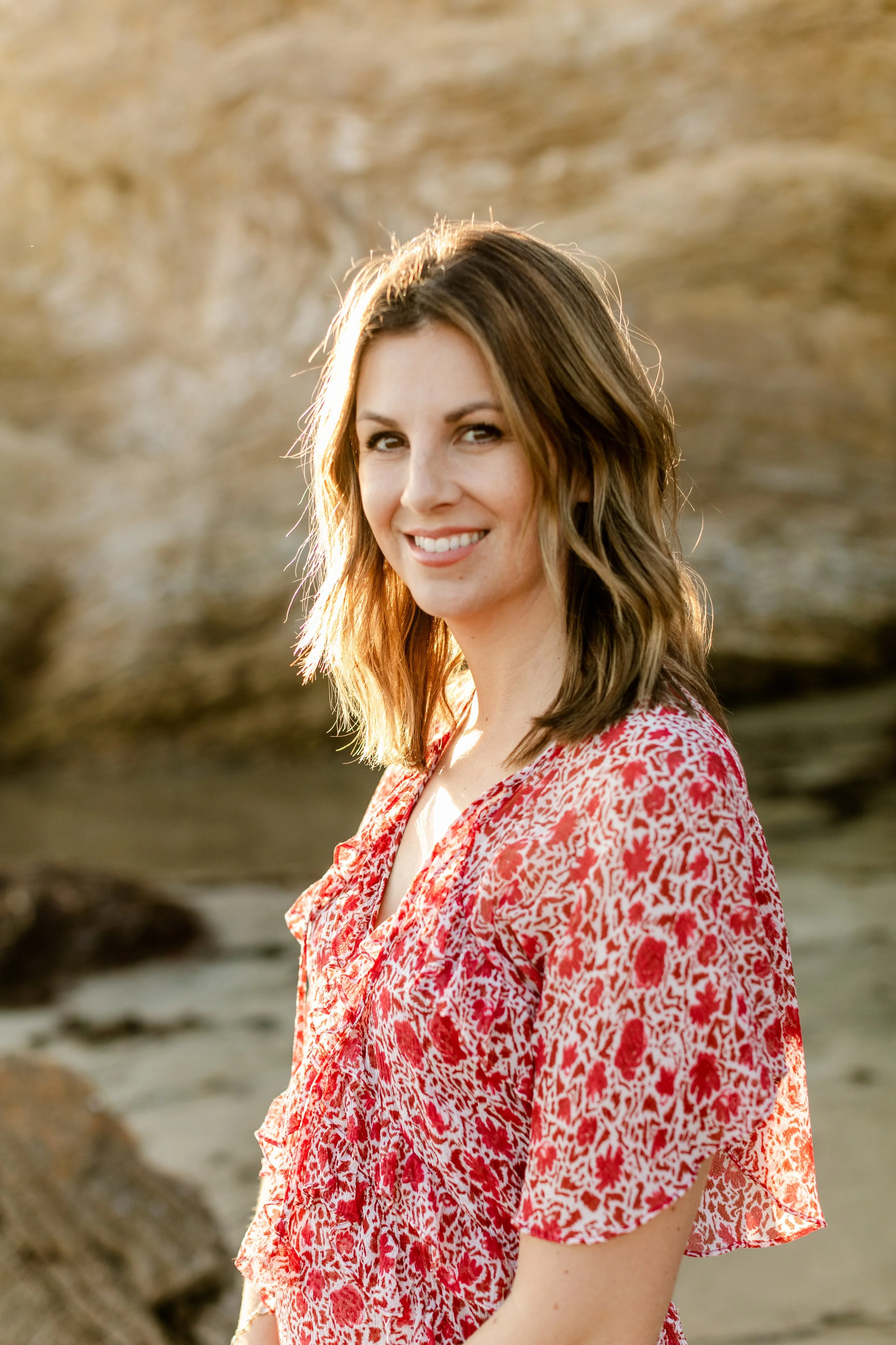 A woman with shoulder-length brown hair, smiling, standing outdoors near rocks, wearing a red and white patterned dress, in warm sunlight.