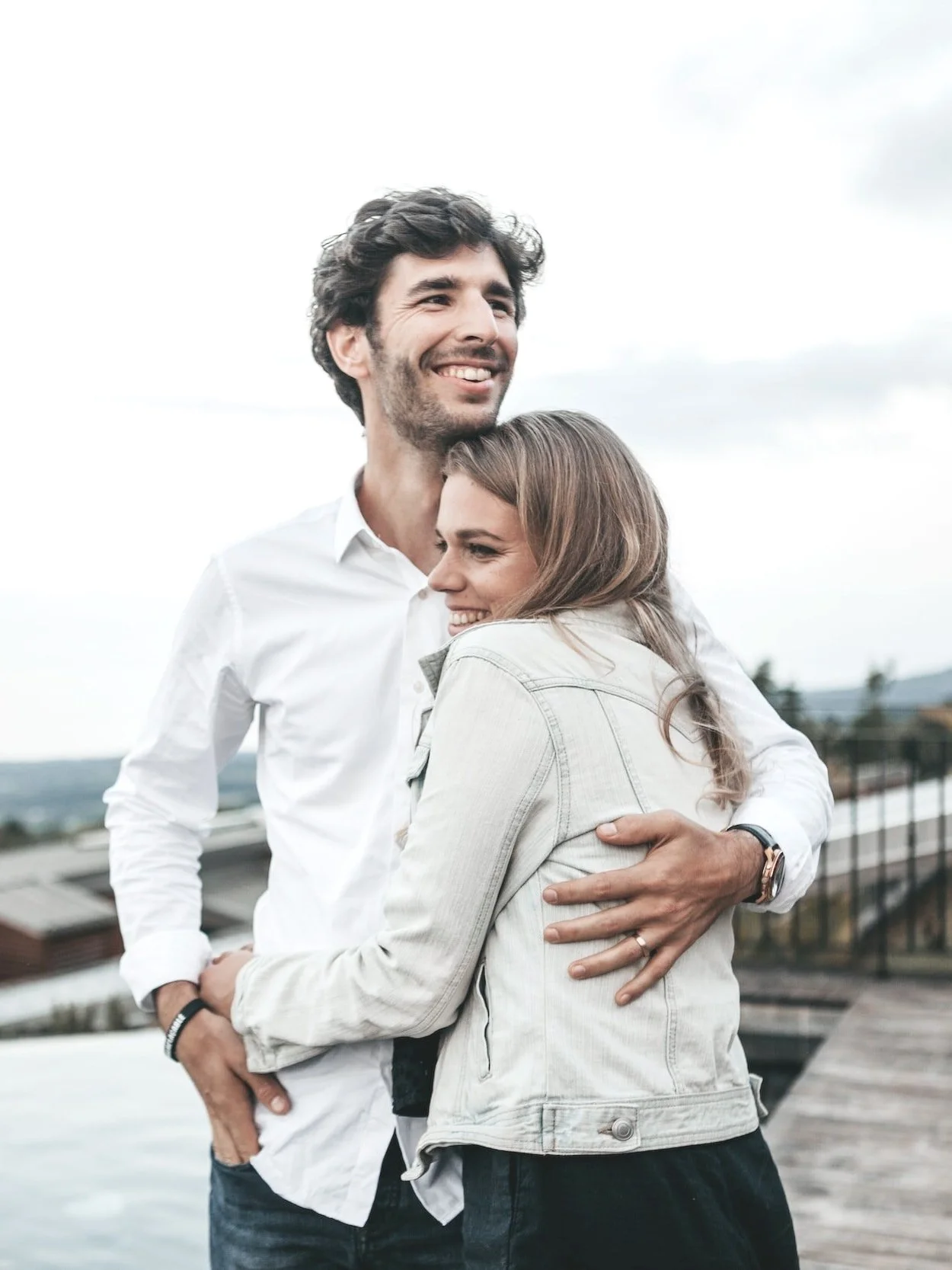 A man and a woman are smiling and hugging outdoors on a cloudy day with a city and water in the background.