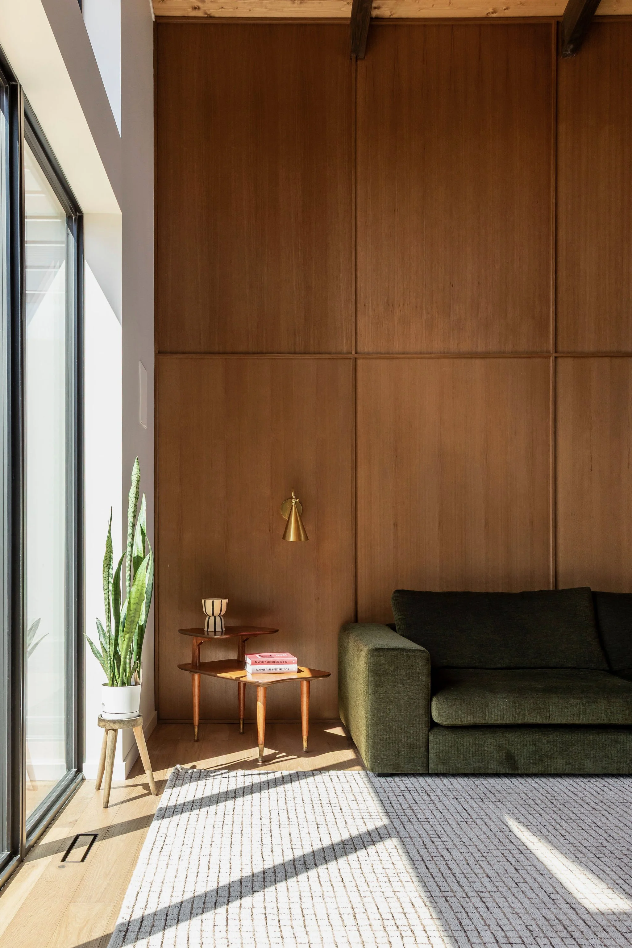 A living room corner featuring a large green sofa, a wooden side table with books and a decorative bowl, a tall potted plant, and a wooden wall with a floating gold lamp. Sunlight streams through a sliding glass door, casting shadows on a light-colored patterned rug.