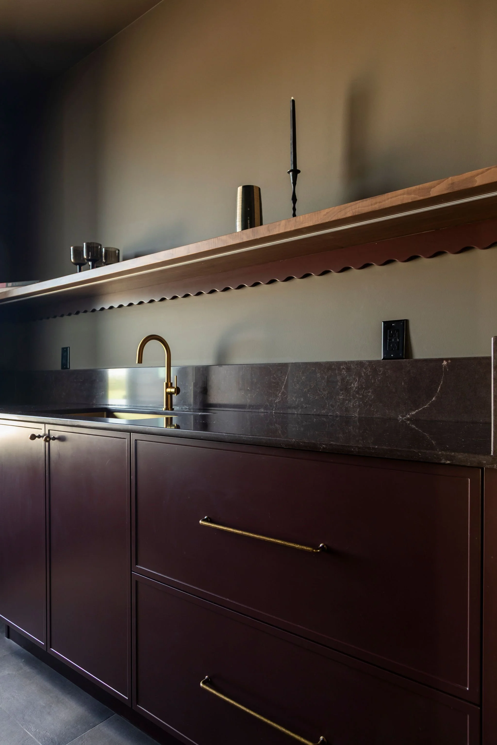 Modern kitchen cabinet with dark wood finish, gold hardware, marble countertop, gold faucet, and a wooden open shelf above with glass cups, a metal tumbler, and a screwdriver.
