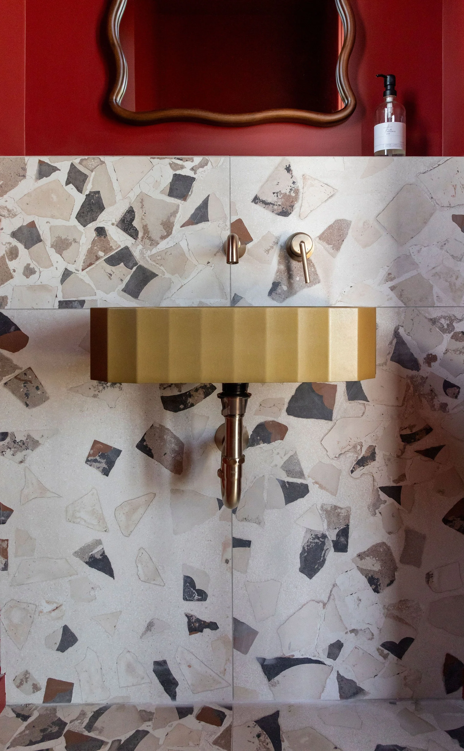 A wall-mounted gold sink with a metallic faucet and a backsplash of mosaic tiles, with a mirror and a soap dispenser on a red-painted wall above it.