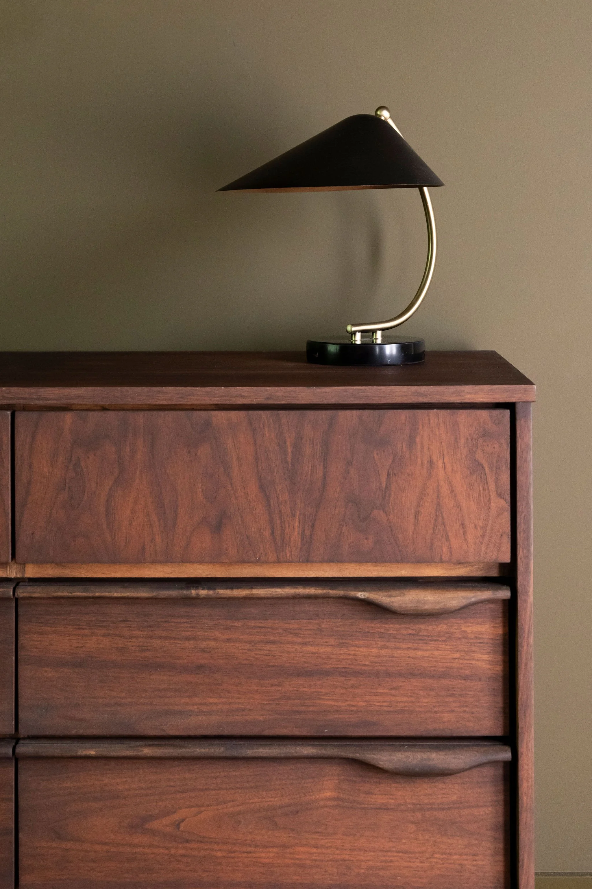 A vintage wooden dresser with a dark brown finish and clean lines, featuring three drawers with curved handles. On top of the dresser is a modern black and brass desk lamp with a curved arm and conical shade, set against a beige wall.