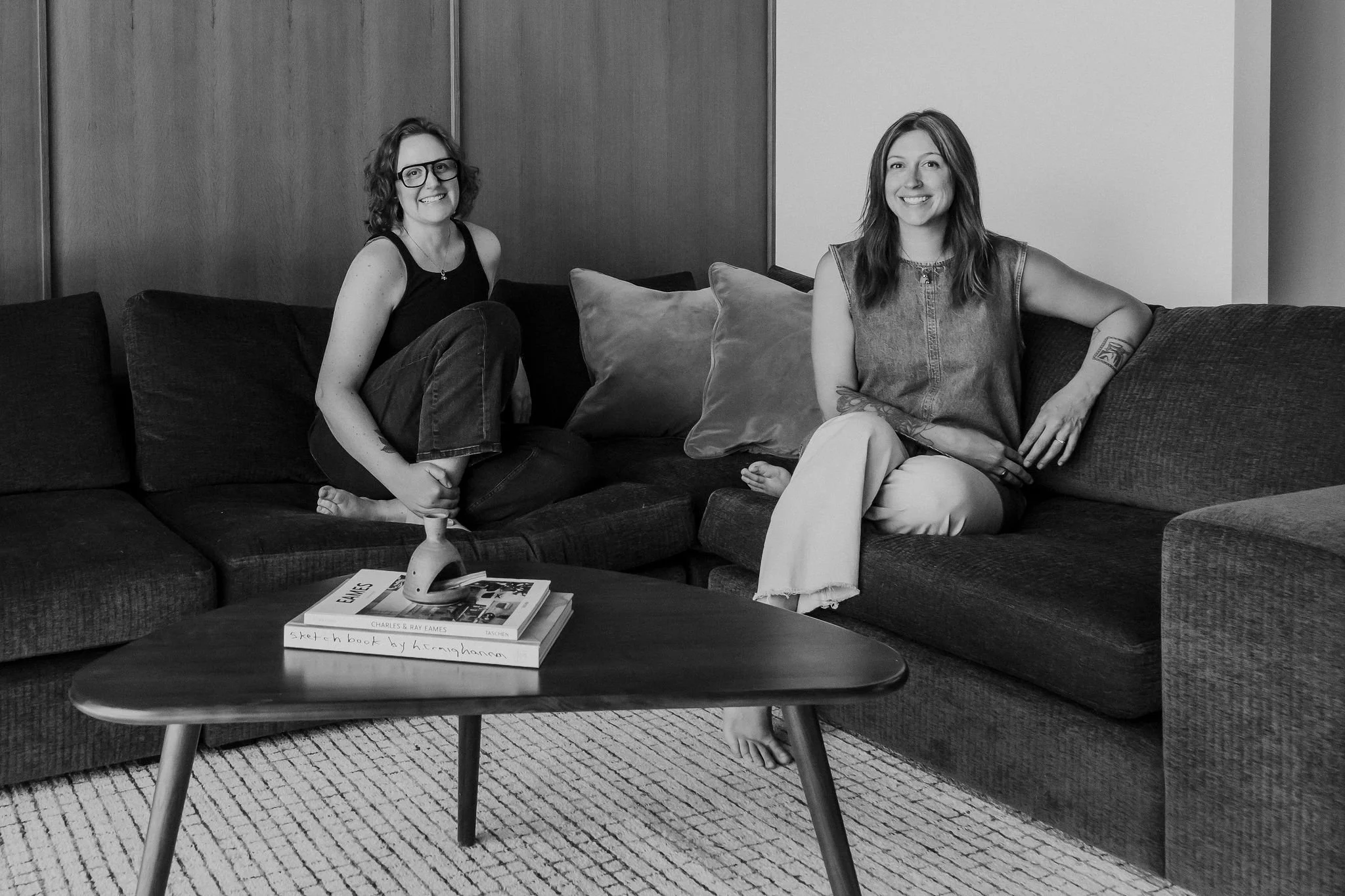 Two women smiling and sitting on a couch in a living room. The woman on the left has glasses and curly hair, the woman on the right has long hair and tattoos on her arms. There is a coffee table with books in front of them.