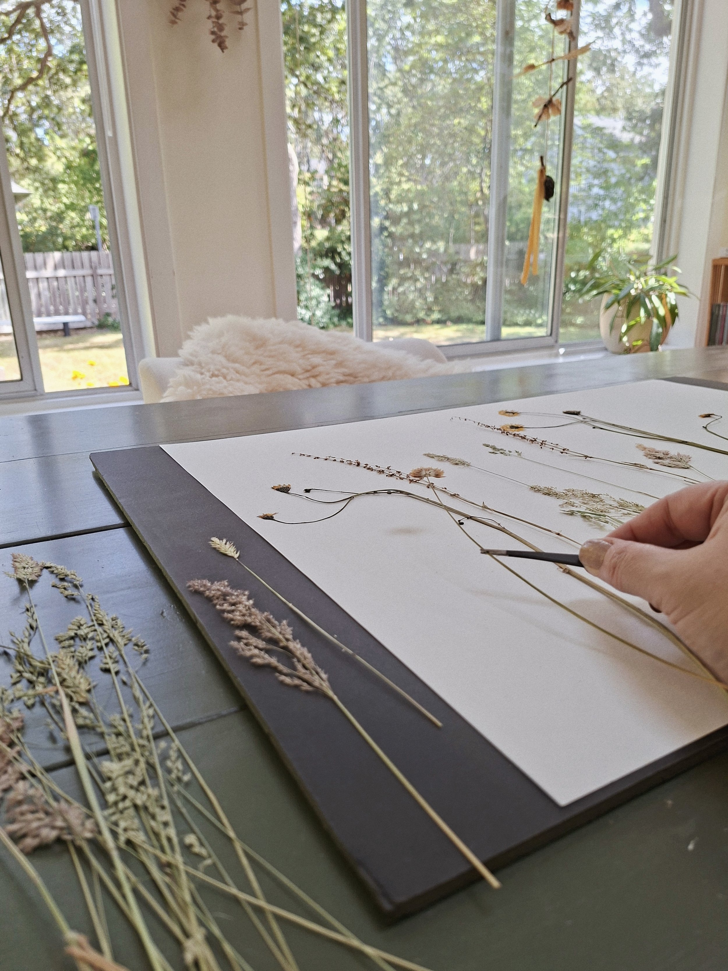 Pressed grasses and wildflowers being arranged by hand into a composition on cream paper at the studio worktable.