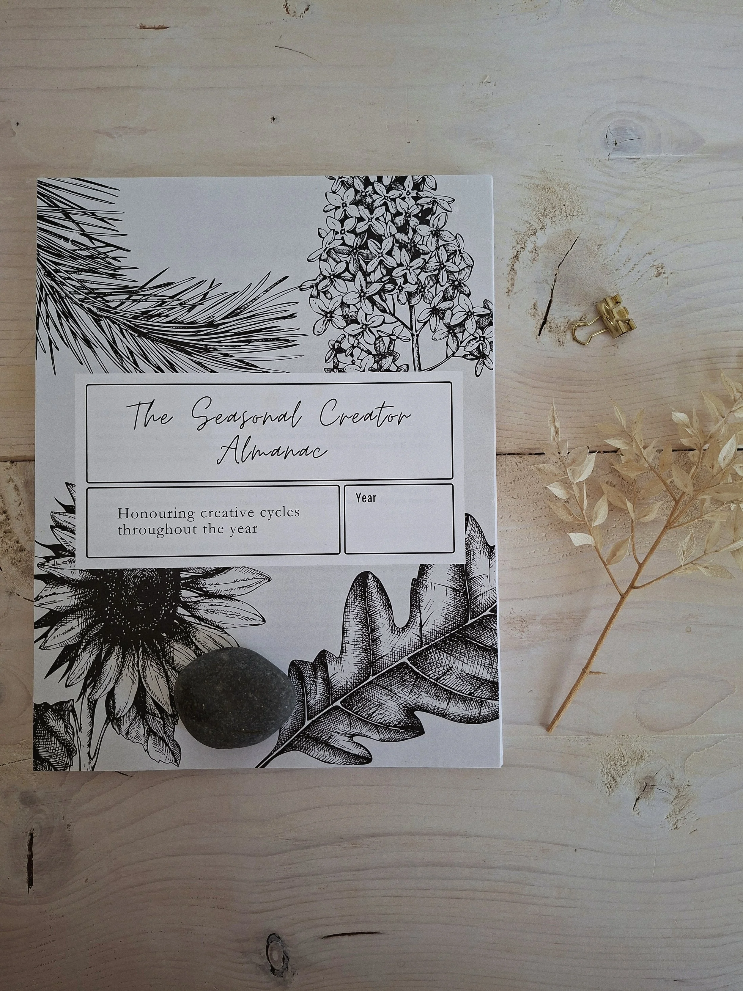 The Seasonal Creator Almanac on a pale wood table with a dried botanical stem and gold clip.