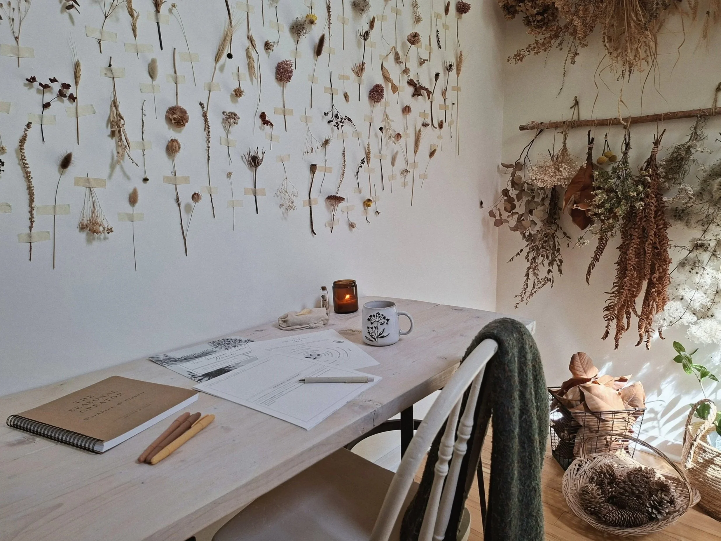 A botanical corner of the studio with The Commons almanac and planner, a candle, mug, pens, seeds, and citrine arranged on a wooden desk.