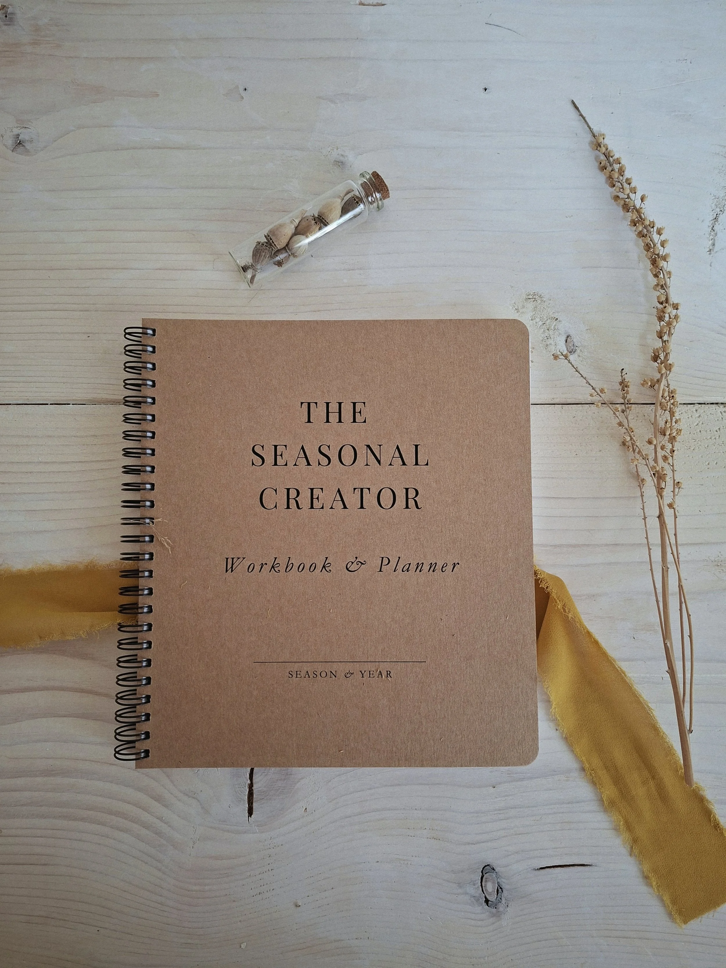 The Seasonal Creator Workbook & Planner on a pale wood table with dried grasses, ochre ribbon, and a small vial of seeds.