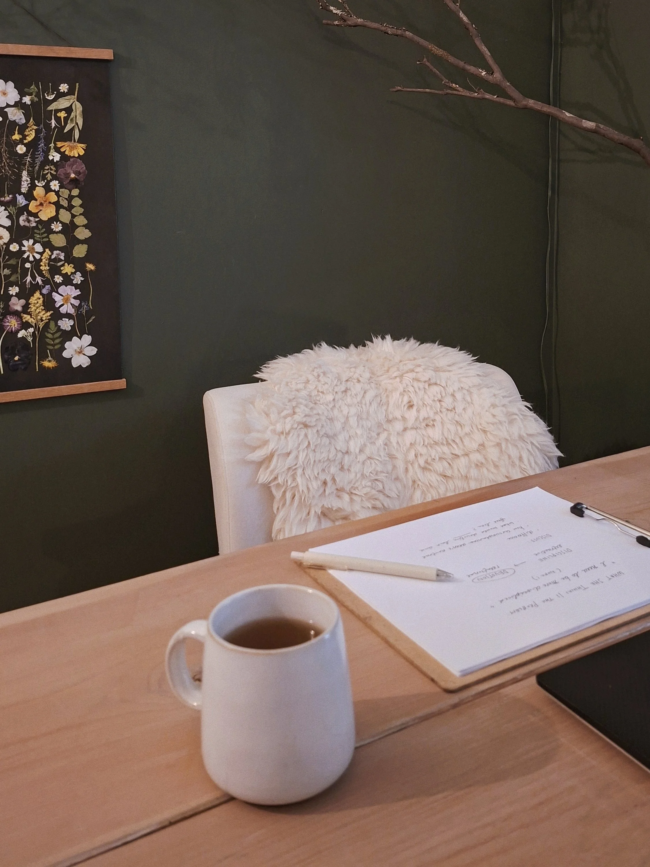 A mug and session notes on a pale wood table, with a pressed-flower wall hanging in the background.