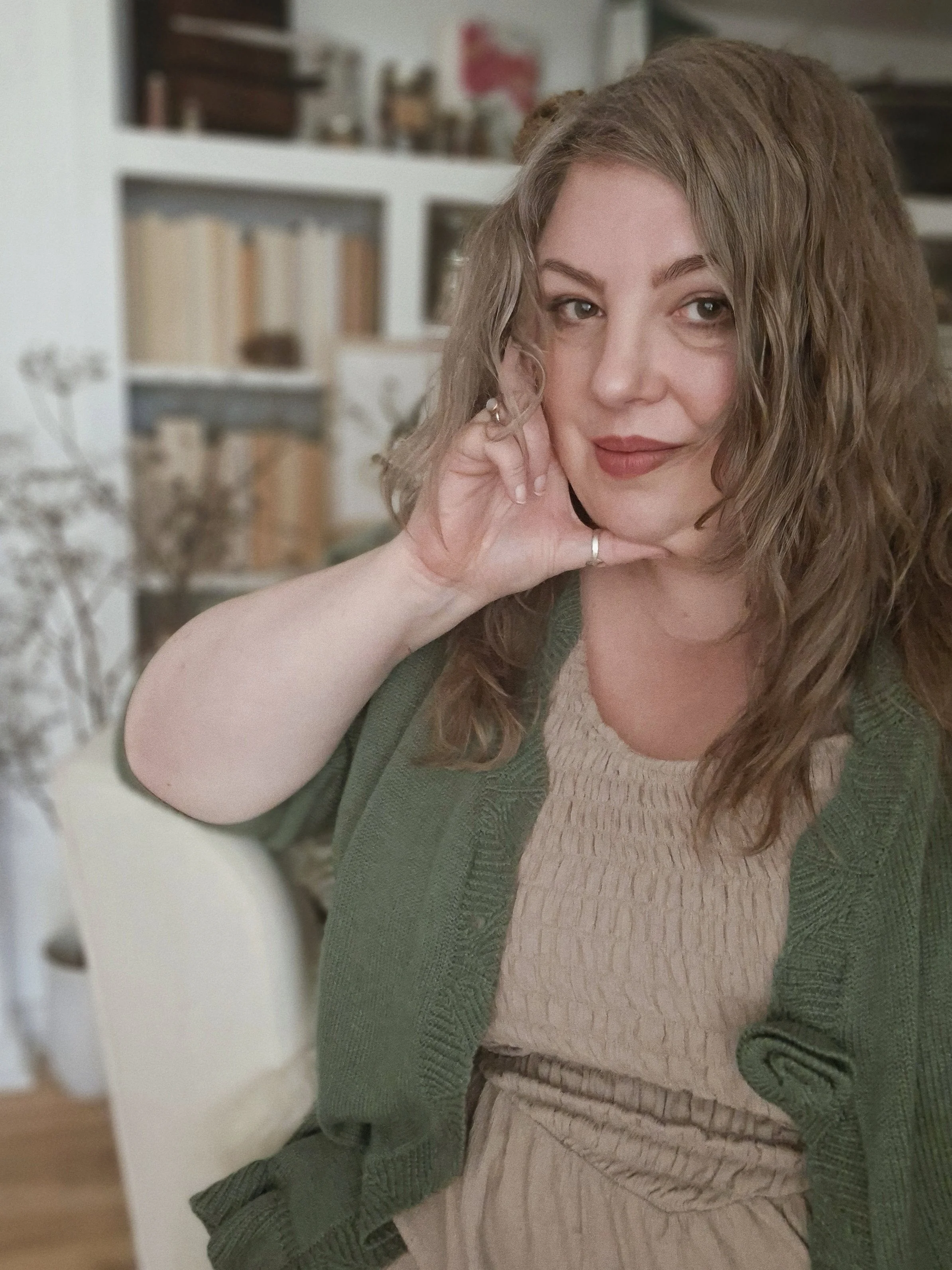 A woman with wavy dark blonde hair and light skin resting her face on her hand, wearing a light taupe-coloured textured dress and a green cardigan, sitting in her studio with bookshelf and decorative items in the background.