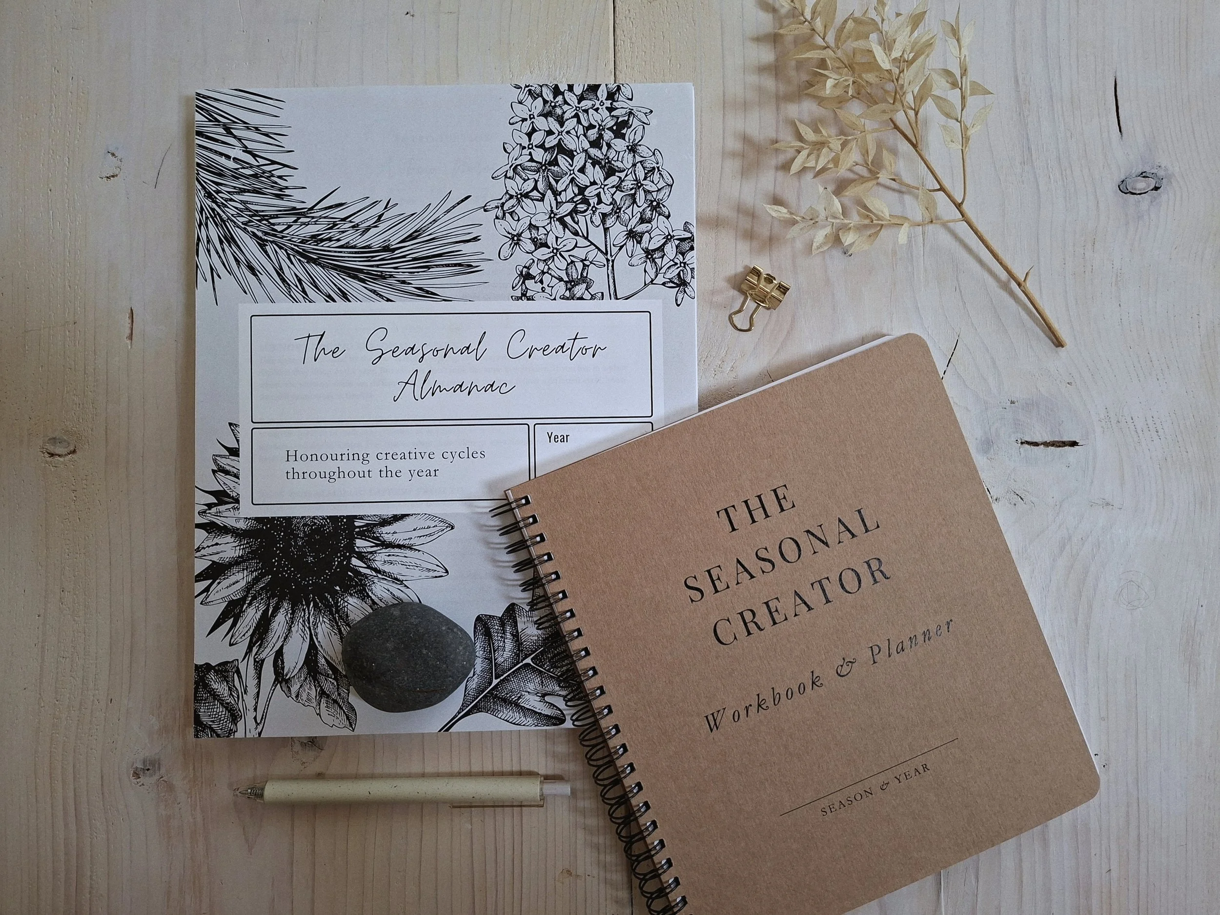 Seasonal Creator Almanac and Workbook & Planner arranged on a pale wood table with a pen, gold clip, and dried botanical stem.