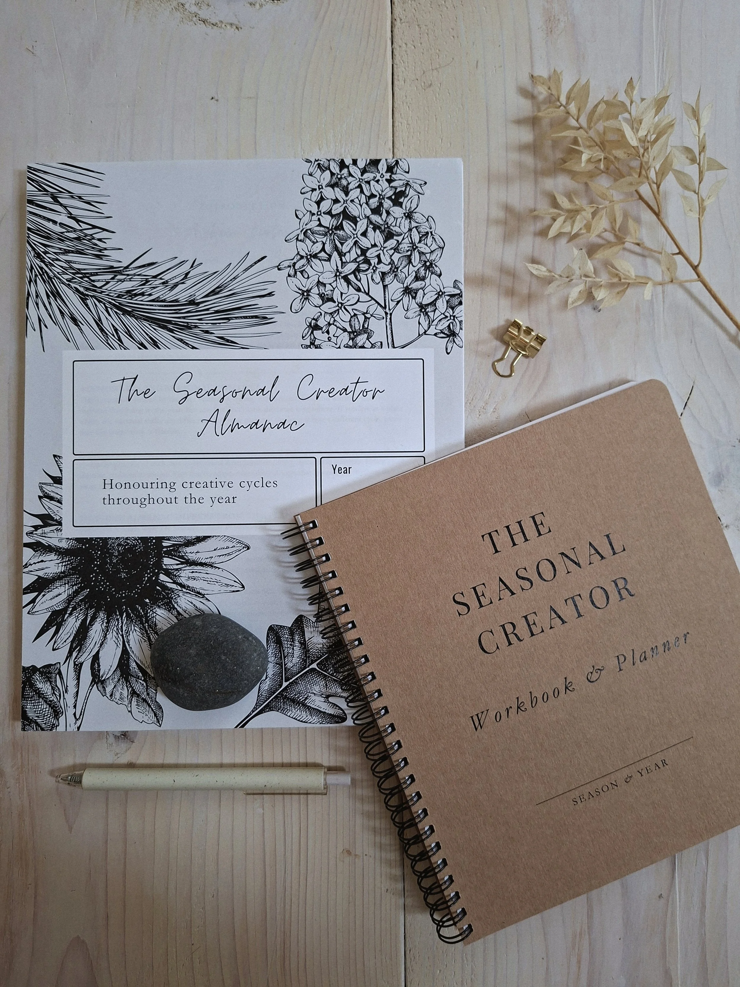Seasonal Creator Almanac and Workbook & Planner arranged on a pale wood table with a pen, gold clip, and dried botanical stem.