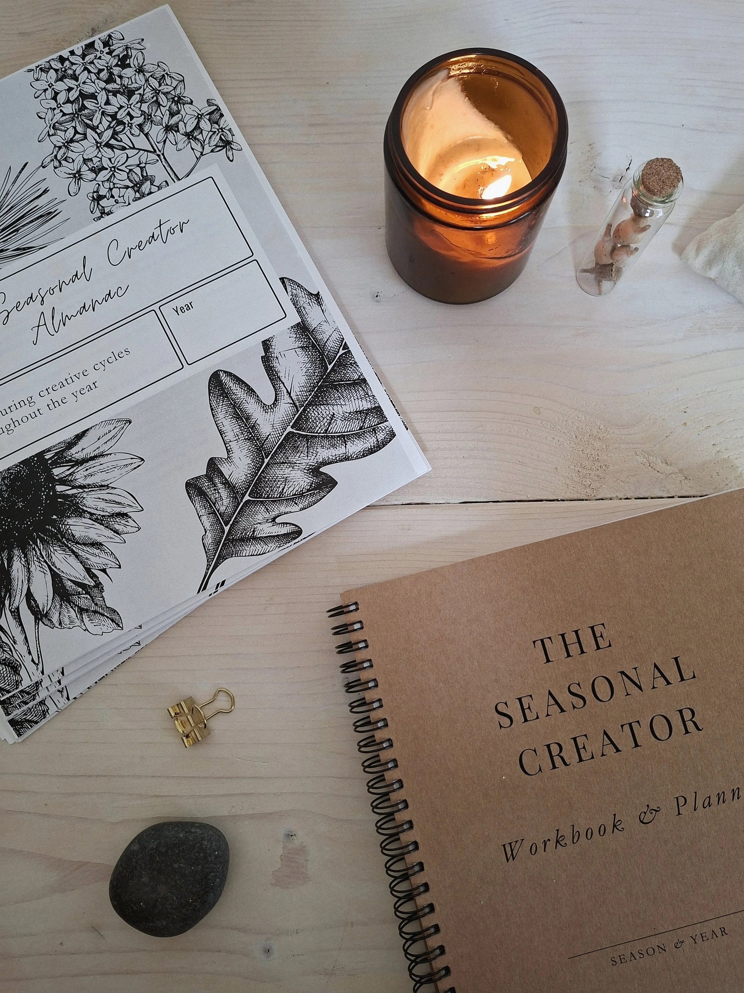 Close-up of The Seasonal Creator Almanac and Workbook & Planner on a pale wood desk with an amber candle, gold clip, and vial of seeds.