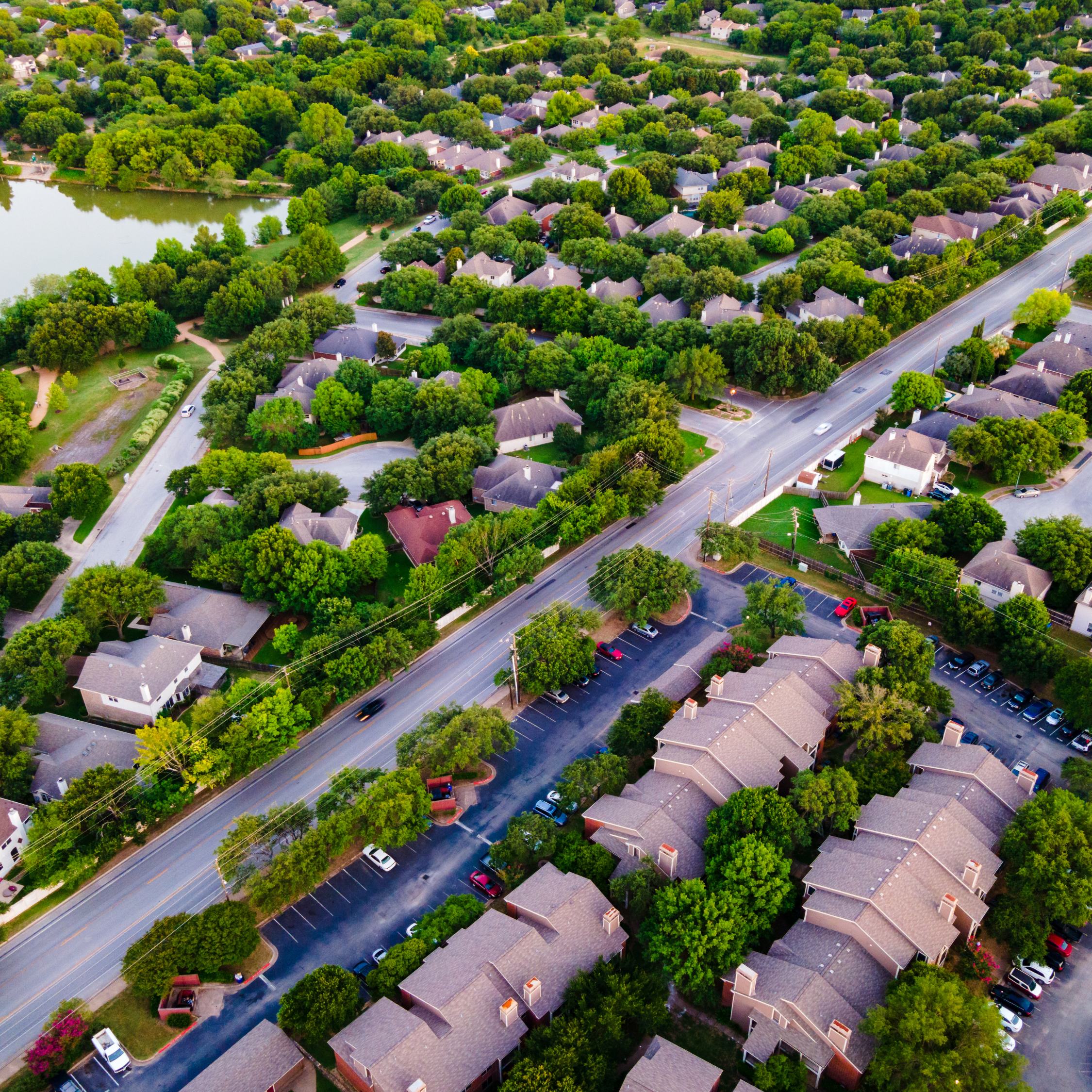 An aerial view of a suburban neighborhood surrounded by trees with a water body on the left side and parking lots on the right.