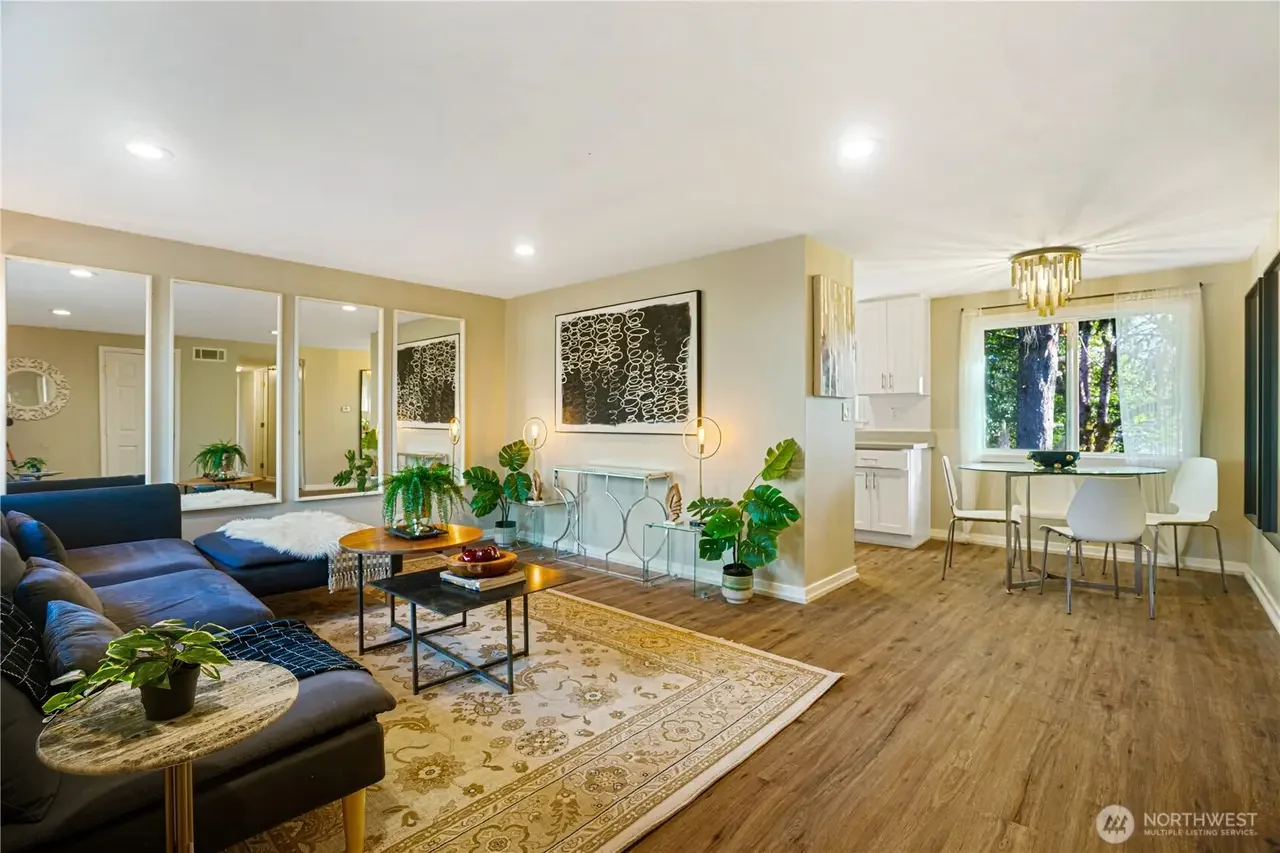 A living room with a dark gray sectional sofa, a patterned area rug, a small round coffee table, houseplants, and wall art. Adjacent to the living room is a dining area with a round table, four white chairs, and a large window with curtains, all illuminated by a modern chandelier.
