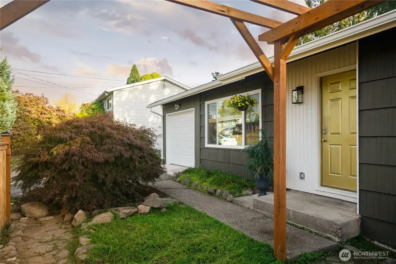 Front yard view of a house with a yellow door, a concrete porch, a small garden area, a hanging flower basket, a bush on the left, and a wood awning frame in the foreground.