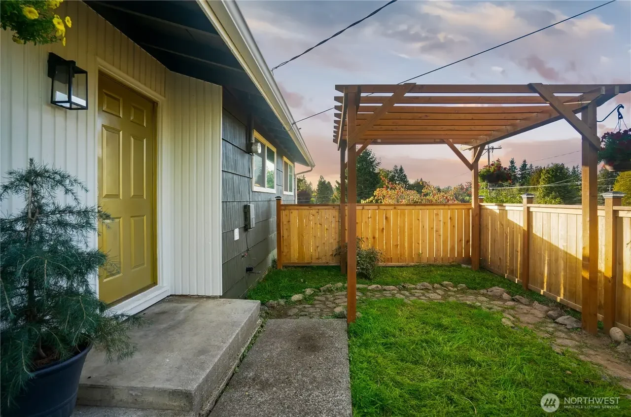 A backyard patio area with a small grassy yard, a wooden fence, and a wooden pergola structure. The house has a yellow door, exterior light, and a potted plant nearby. The sky is partly cloudy during sunset.