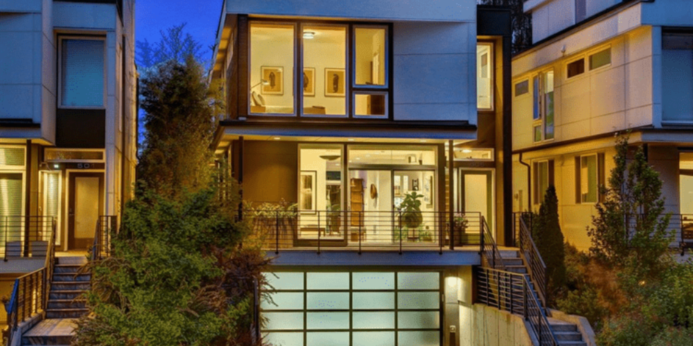 Modern multi-story house with large windows, surrounded by trees and greenery, illuminated at night.