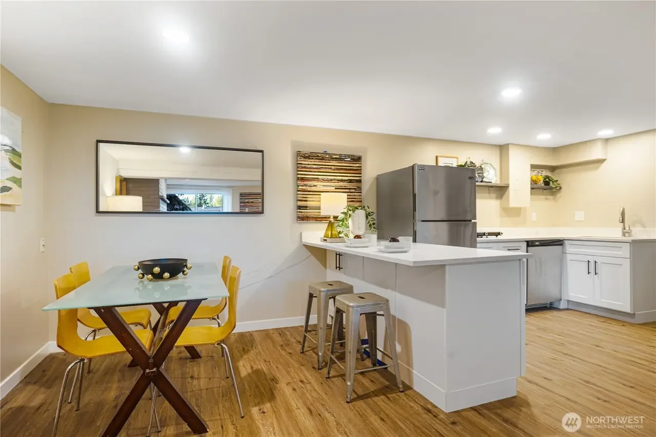 Open kitchen and dining area with a rectangular table and yellow chairs, stainless steel refrigerator, white cabinets, wooden flooring, and decorative wall art.