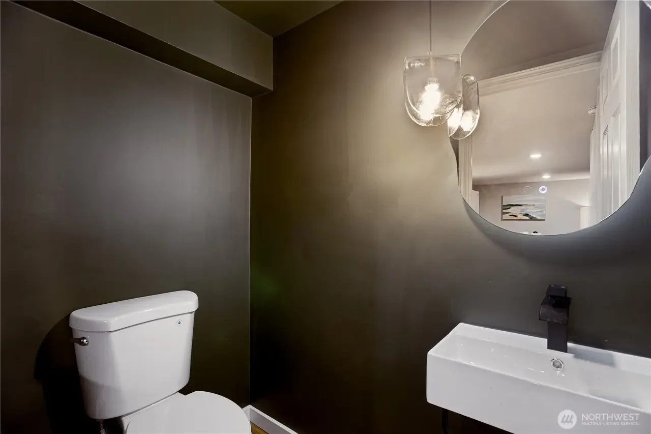 A modern bathroom with a white toilet and a small rectangular sink with a black faucet, a round mirror with a curved top, and glass pendant lights hanging from the ceiling. The walls are painted dark, and a part of an art piece is visible in the refl