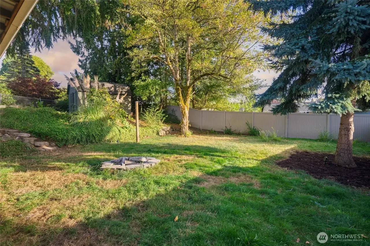 A backyard with green grass, trees, a shed, and a white fence. There are some flower beds and a small fire pit.