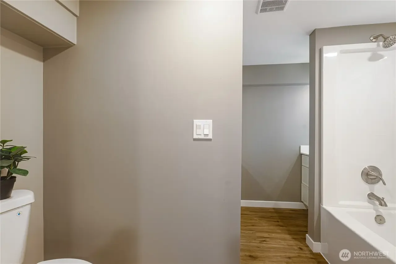 Interior view of a bathroom showing a toilet with a potted plant on top and part of a bathtub with a showerhead, wall switches, and hardwood flooring.