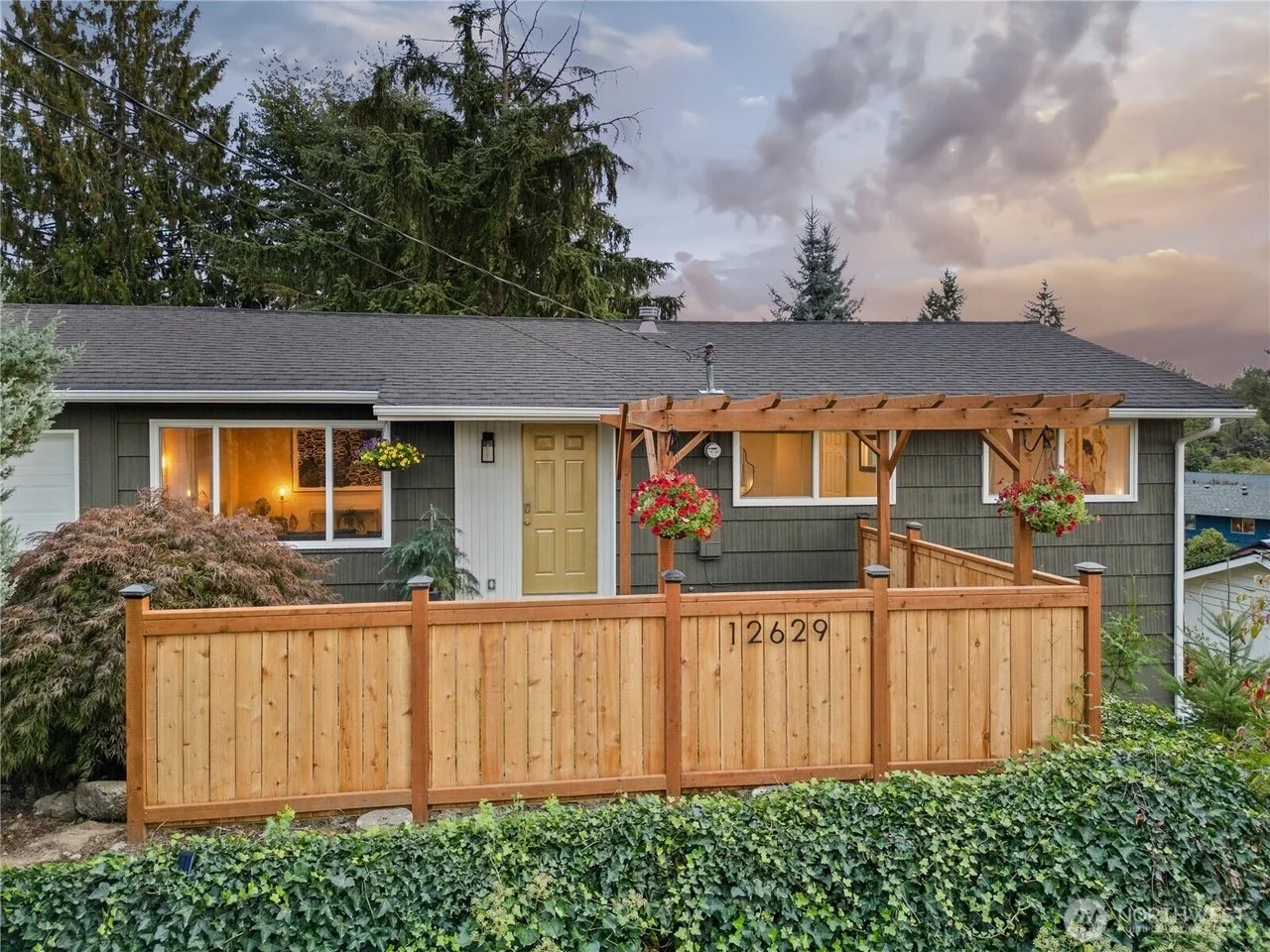 Front view of a house with a green exterior and a wooden fence with gates, featuring hanging flower baskets and a small pergola with hanging flower baskets, at sunset or early evening.